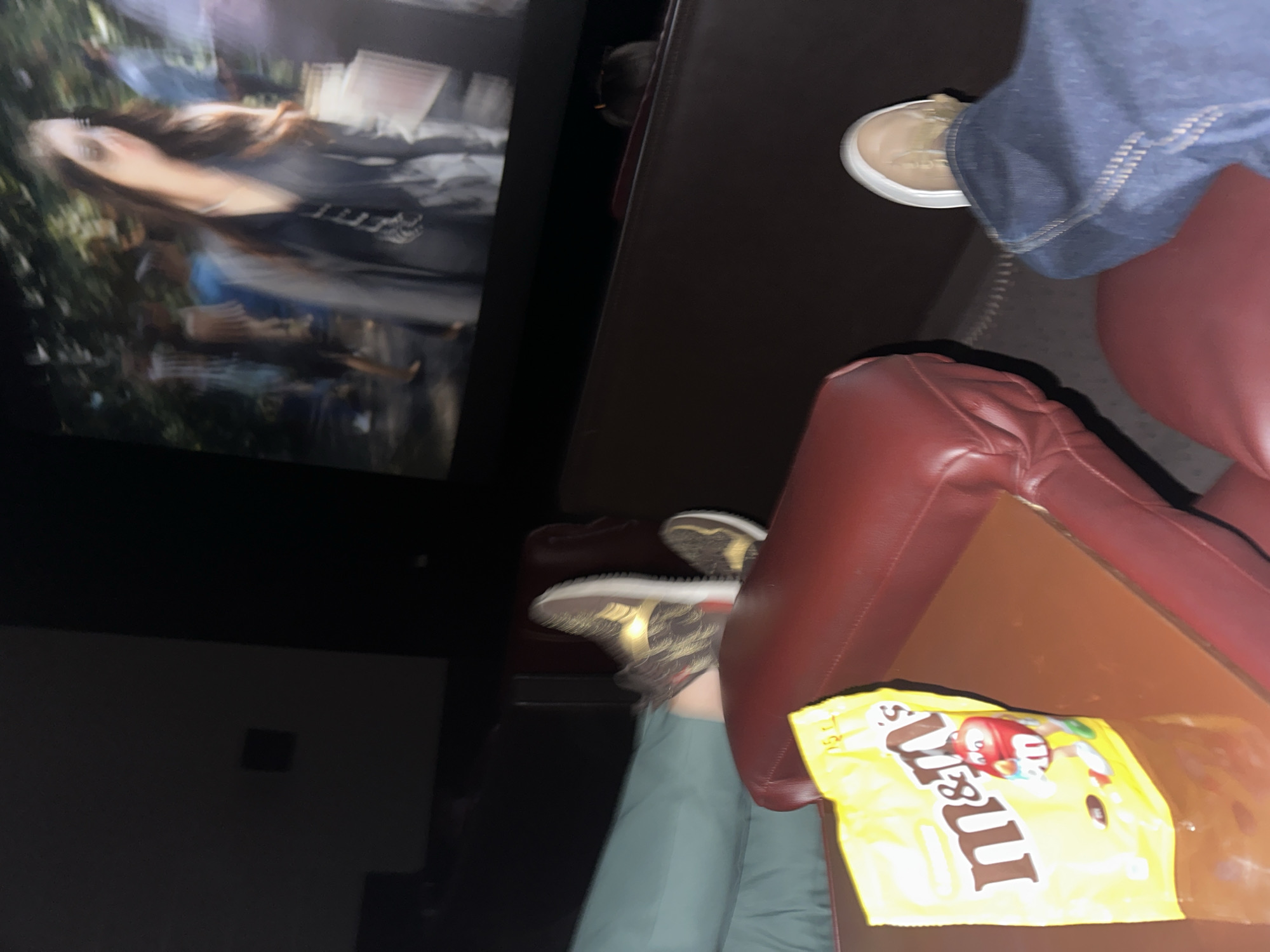 The image captures a scene inside a movie theater. A person is sitting in a red leather seat, their legs crossed, with a bag of M&M's on the armrest. Another person's legs and shoes are visible in the adjacent seat. The movie screen in the background shows a blurred image of a woman. The location is likely in Yerevan, Armenia, but this cannot be definitively confirmed from the image alone. The lighting is dim, typical of a movie theater during a screening.
