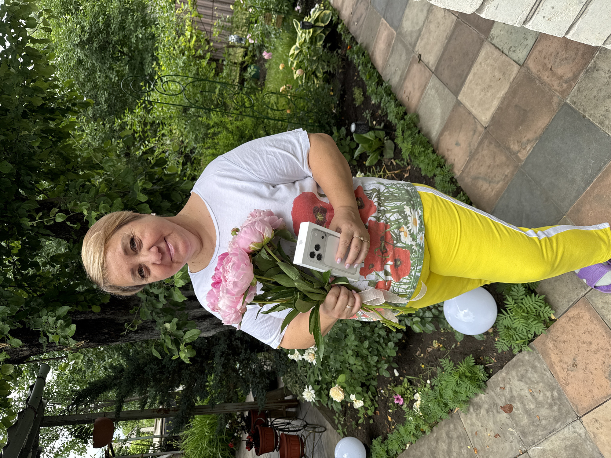 In the city of Nizhne-Chufichevo, Russia, a woman with short blonde hair stands in a garden setting, holding a bouquet of pink peonies and a silver iPhone 14 Pro Max in its box. She is wearing a white t-shirt with a floral and poppy print and bright yellow sweatpants with a white stripe. The garden features lush green trees and bushes, with a paved walkway made of square tiles in earthy tones. Several white spherical garden lights are visible, along with potted plants and decorative garden features. The woman is smiling and looking at the camera.