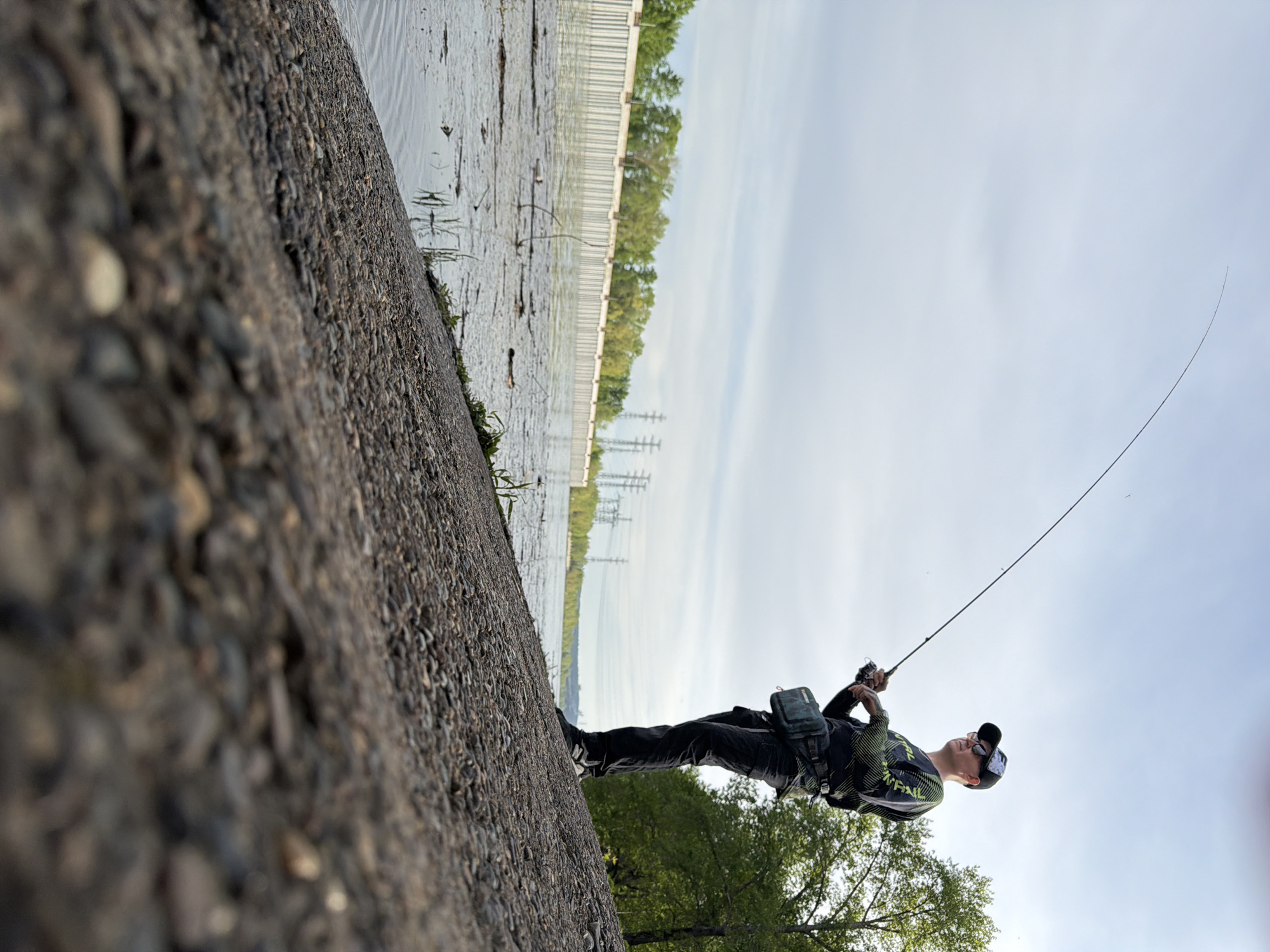 A man is fishing at a body of water in what appears to be the city of Chaikovsky, Russia. The shoreline where the man stands is composed of a steep slope of small rocks and debris. The water is to his left, and across the water are trees and utility towers. The man wears dark pants, a patterned shirt, a dark cap, and a shoulder bag. He is holding a fishing rod, which extends upwards into the sky. The sky is partially cloudy.