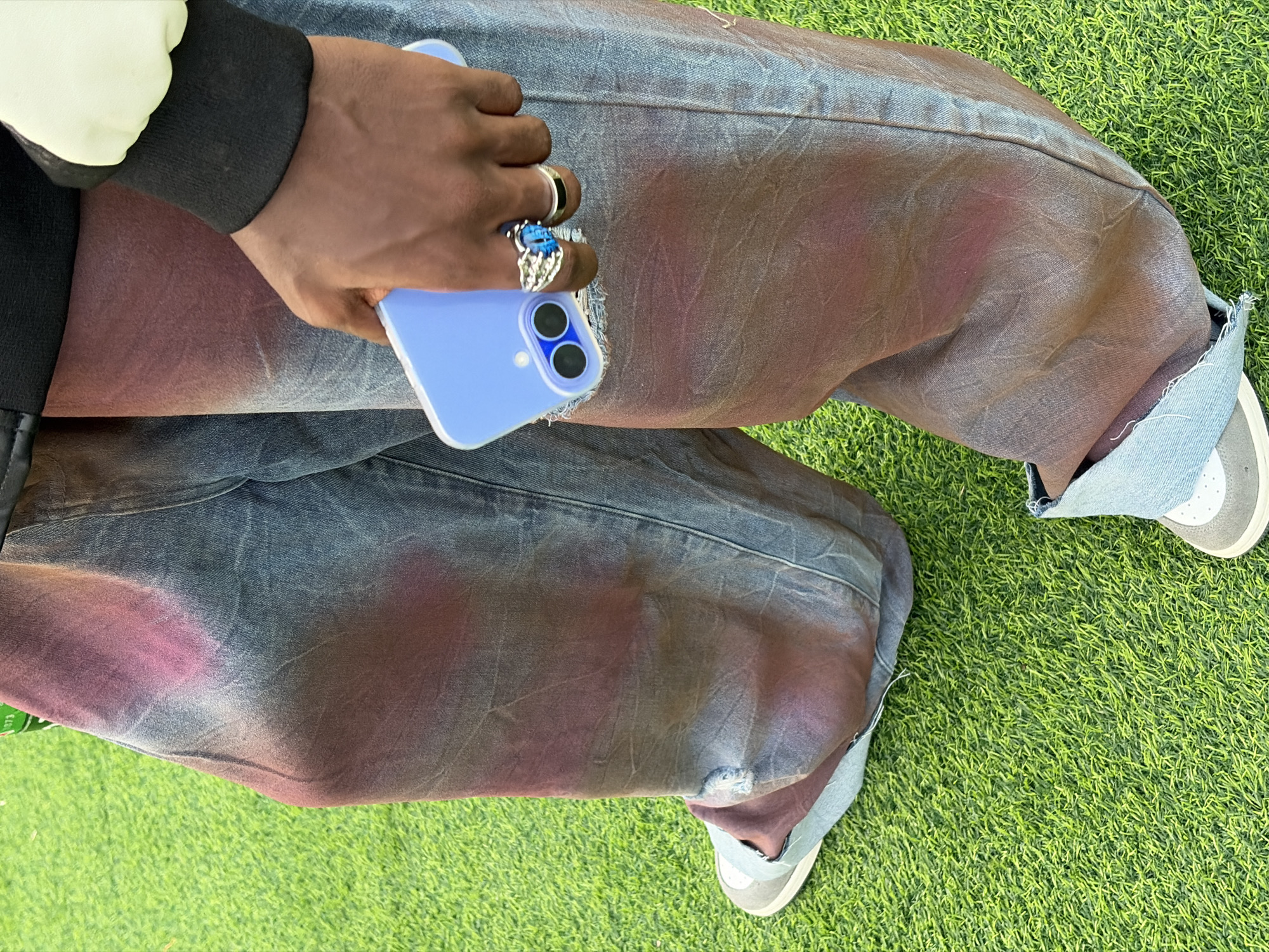 A medium shot from an elevated perspective shows a person seated on artificial turf. The person is holding a light blue cell phone in their left hand, which also has three rings. The person is wearing jeans, which have been dyed blue, brown, and purple and rolled to expose blue linings on the bottom. The person wears a black and white jacket and light-colored sneakers. The scene is on green artificial turf. There is no evidence in the image to suggest this is Lagos, Nigeria.