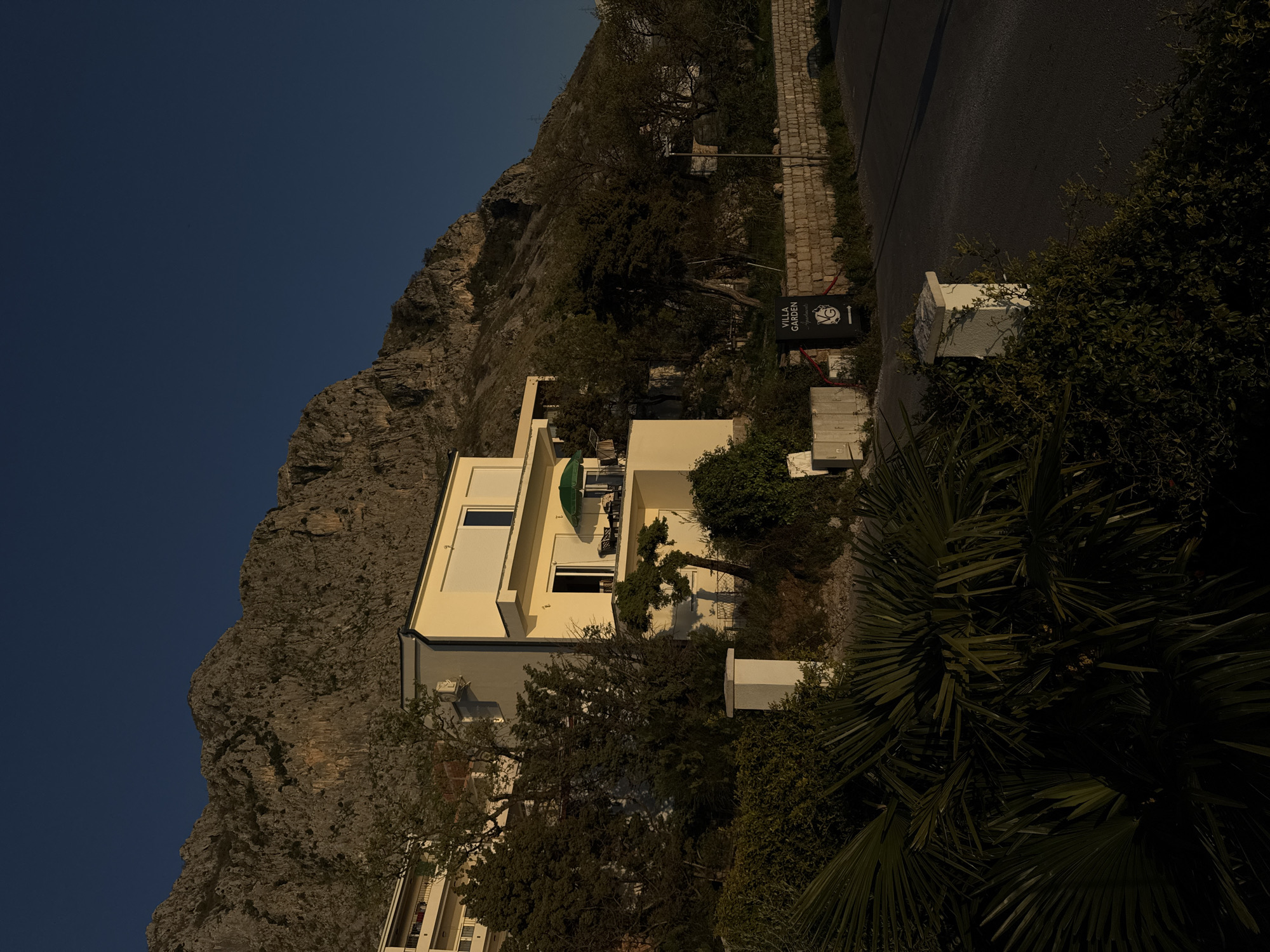 A medium, outdoor shot, angled to the left, showcases a multi-story house, set against the backdrop of a rocky mountain and a clear blue sky. The house is cream-colored, featuring multiple windows and a balcony with a green umbrella. The mountain, with its rough texture and varying shades of gray and brown, occupies the left side of the frame. The sky, a deep shade of blue, provides a stark contrast to the mountain. To the right of the house, a road stretches into the distance. Lush vegetation, including palm trees, bushes, and other greenery, is scattered around the house and along the road, partially obscuring the view. A sign reading "Villa Garden Apartments" is visible near the road. The lighting suggests either early morning or late afternoon, casting long shadows and creating a sense of depth. The scene captures a serene setting, blending architecture with the natural landscape in Blizikuće, Montenegro. There are no people shown in this image.
