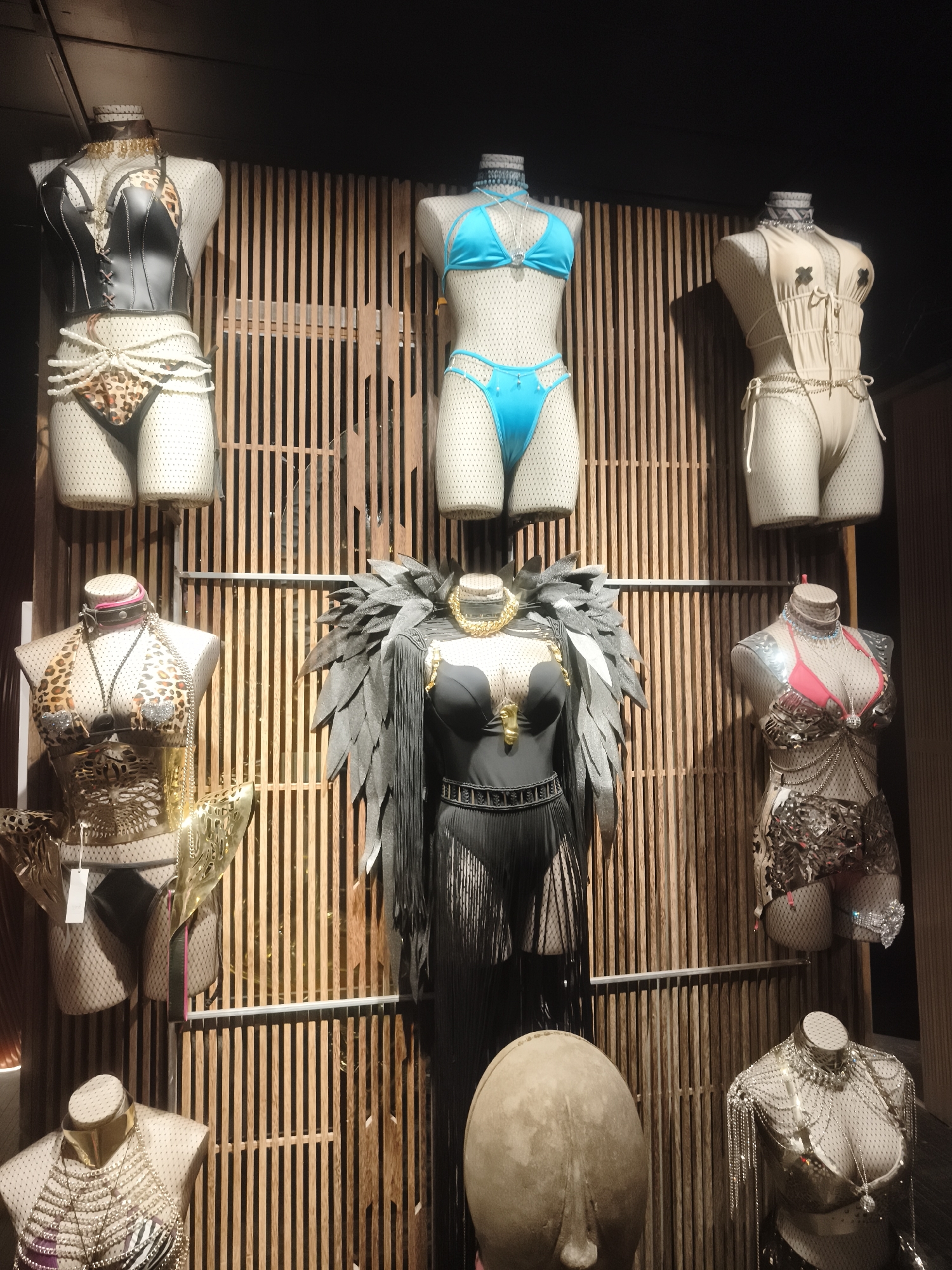 A retail display in Baan Mae Nam, Thailand, features multiple mannequins adorned with elaborate and suggestive outfits. The display is arranged against a backdrop of vertical wooden slats. In the top row, from left to right, are three mannequins. The leftmost mannequin wears a black leather-like corset with a leopard print bikini bottom, accented with pearl-like beading around the waist. The middle mannequin is dressed in a bright turquoise bikini set, including a triangle top and low-rise bottoms with cutouts. The rightmost mannequin is outfitted in a beige bikini with black pasties, a cinched waist, and drawstring details.
In the middle row, also from left to right, are three more mannequins. The leftmost mannequin sports a leopard print halter top bikini with a caged-like gold metallic skirt accessory. The central mannequin is dramatic, wearing a black outfit featuring large, dark feathery wings, a plunging neckline, and a pleated skirt. The rightmost mannequin in this row wears a pink bikini top paired with a metallic, scale-like armor piece extending from the chest down to the hips.
The bottom row consists of a large, abstract, egg-shaped sculpture in the center. To the left of the sculpture, a mannequin wears a gold collar and a multi-layered necklace with chain and bead embellishments. To the right of the sculpture, another mannequin displays a silver chainmail-like bra and skirt set, with a pendant necklace. The overall presentation suggests a boutique or specialty store specializing in avant-garde or performance-wear.