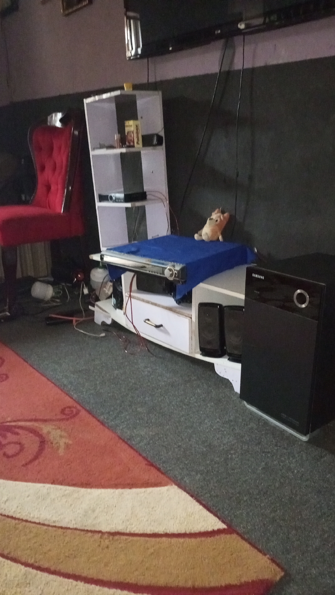 The image shows a living room in Unknown, Unknown. A red, high-backed chair sits to the left of a white shelving unit that contains a DVD player and a box of Pringles. Wires are tangled on the floor around these items. A white TV stand holds a covered device with a blue cloth. On top of the blue cloth is a stuffed animal. A black Samsung speaker is positioned to the right of the TV stand. A large rug with red, white, and brown circular patterns is in the foreground. A flat screen TV is mounted high on the wall. The walls are a dark grey color.