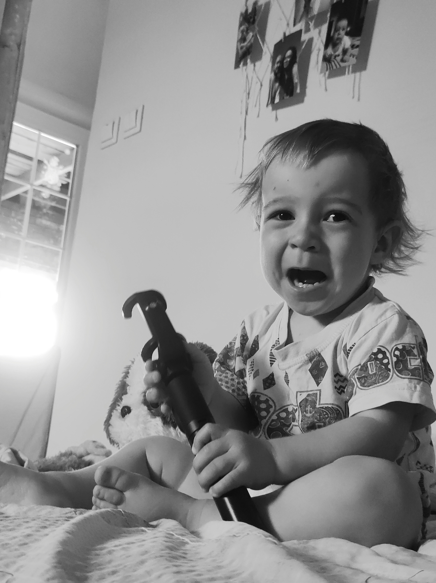 A black and white medium shot shows a young child sitting cross-legged on a bed, holding a black object that resembles a pump or tool. The child is looking directly at the camera with an open mouth, appearing upset or crying. To the child's left is a plush toy animal, partially obscured by the object. In the background, there is a window on the left and several photographs hanging on the wall above the child's head. The bed is covered with a textured blanket. The location is Unknown, Unknown.