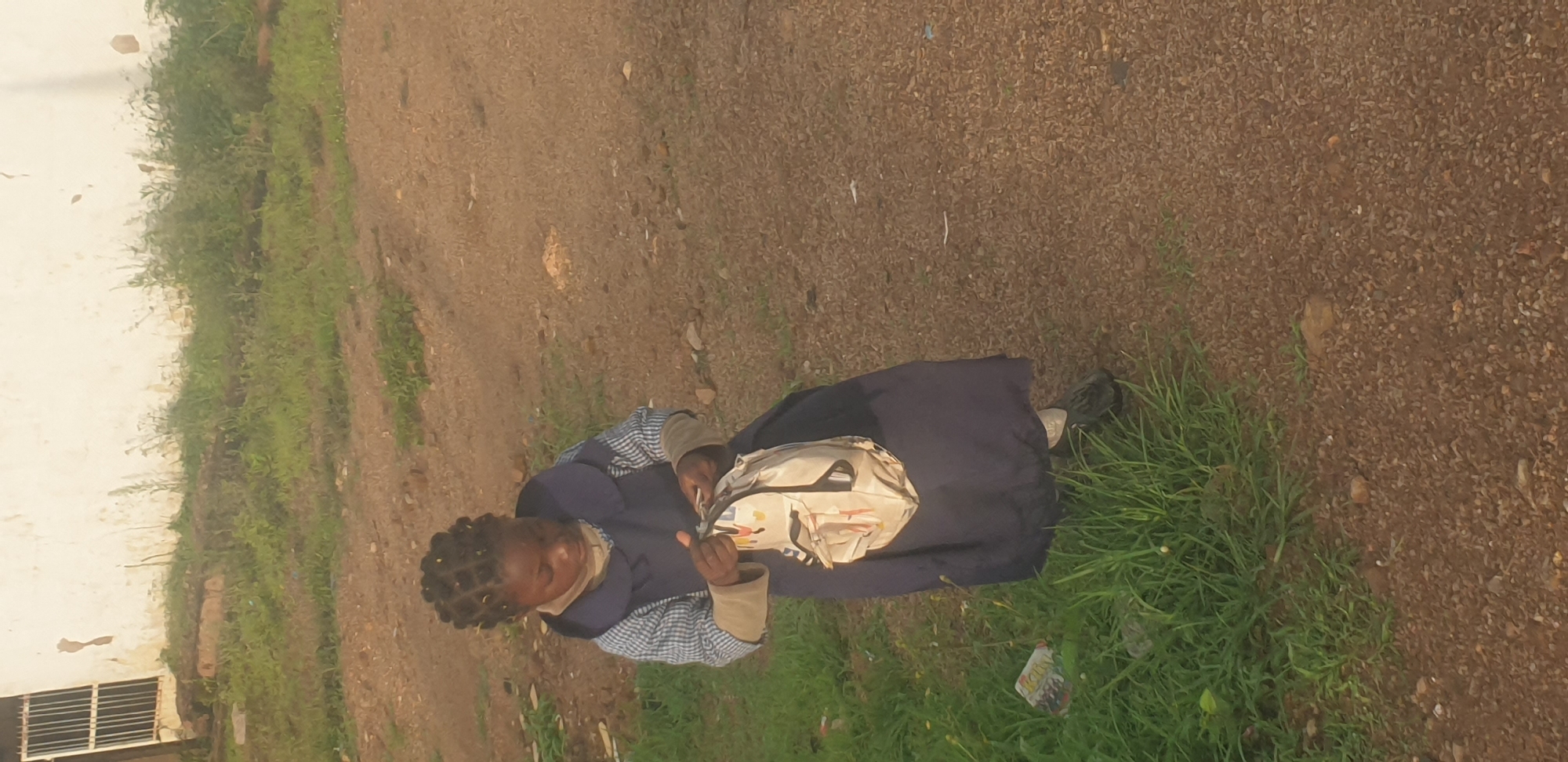 In the city of Jos, Nigeria, a young girl with braided hair, wearing a school uniform consisting of a blue pinafore over a blue and white striped shirt, stands outdoors. She is holding a light-colored backpack with a patterned design. The ground around her is a mix of dirt, gravel, and patches of green grass. In the background, a weathered, light-colored building with barred windows is visible, along with more vegetation. The scene appears to be a typical street or schoolyard environment.