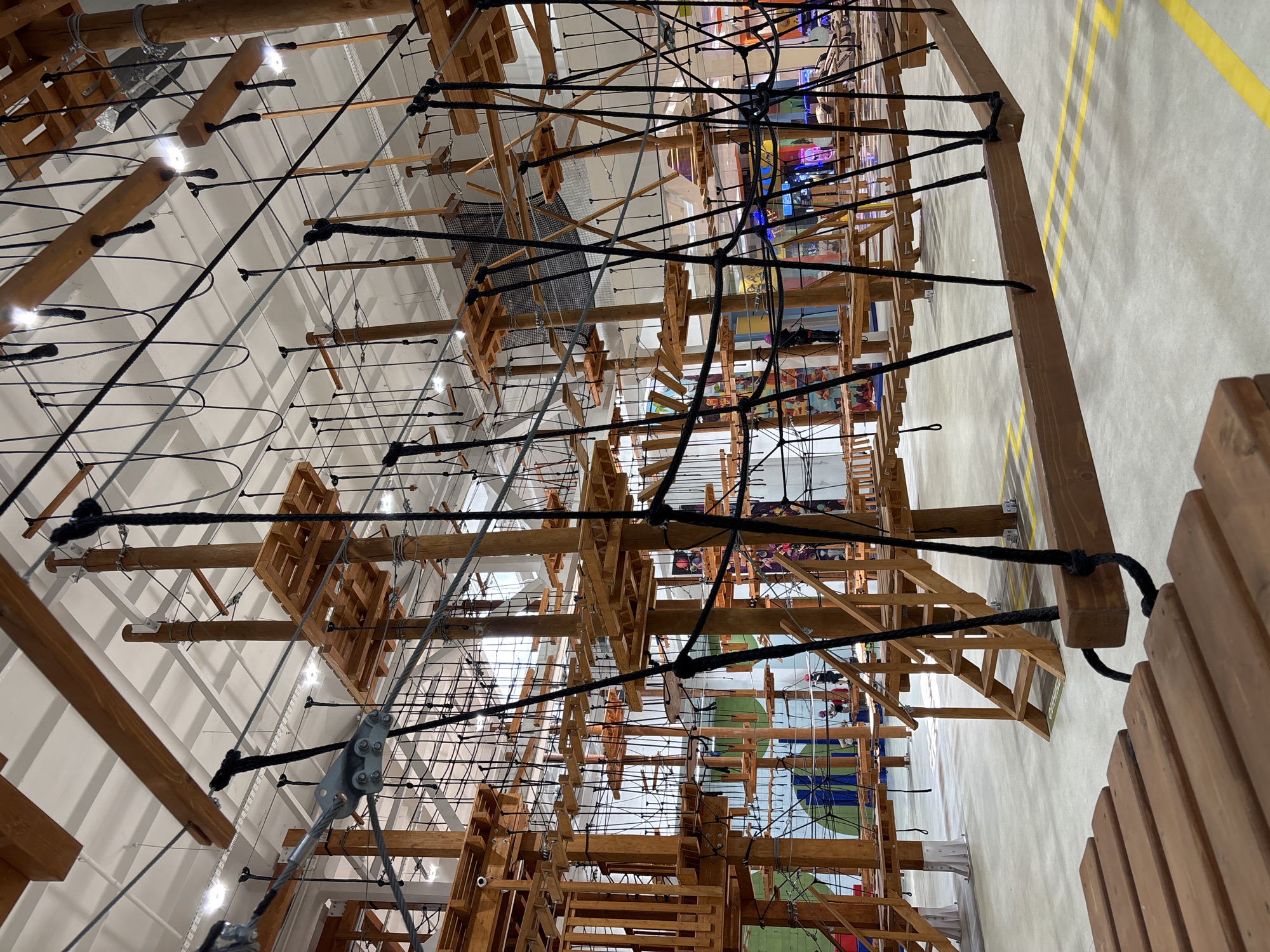 The image shows a high ropes course inside an indoor recreational facility. The structure is made primarily of wood and rope, with multiple levels of platforms, bridges, and obstacles. Numerous thick ropes are suspended from the ceiling and connect various parts of the course. Wooden beams and planks form walkways and supports. The floor is a smooth, light-colored concrete with yellow safety lines. In the background, there are glimpses of colorful decorations and lighting, suggesting an entertainment venue. A few people are visible on the ropes course, appearing to navigate the challenges. One person in a red helmet is standing on a platform. Another person in a pink helmet is standing further back on a bridge. The lighting in the facility is bright, with visible fluorescent lights in the ceiling. There is no indication of the city of Orenburg, Russia in the image.