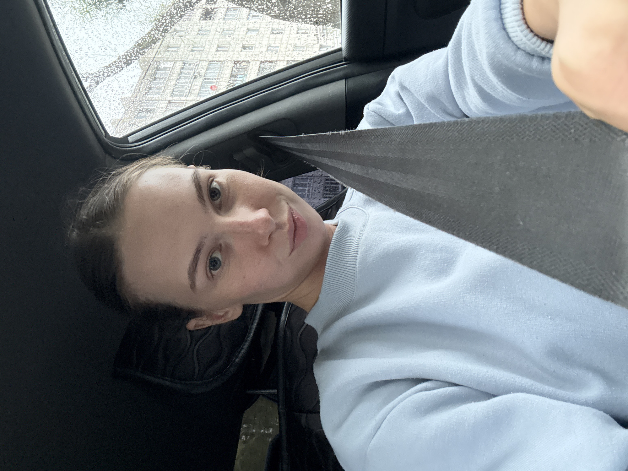 A young woman with blonde hair pulled back into a bun is seated in the driver's seat of a car, looking at the camera. She is wearing a light blue sweatshirt. A gray seatbelt is diagonally across her chest. Rain streaks are visible on the car window, obscuring the view of a building outside. The scene appears to be in the city of Perm, Russia.