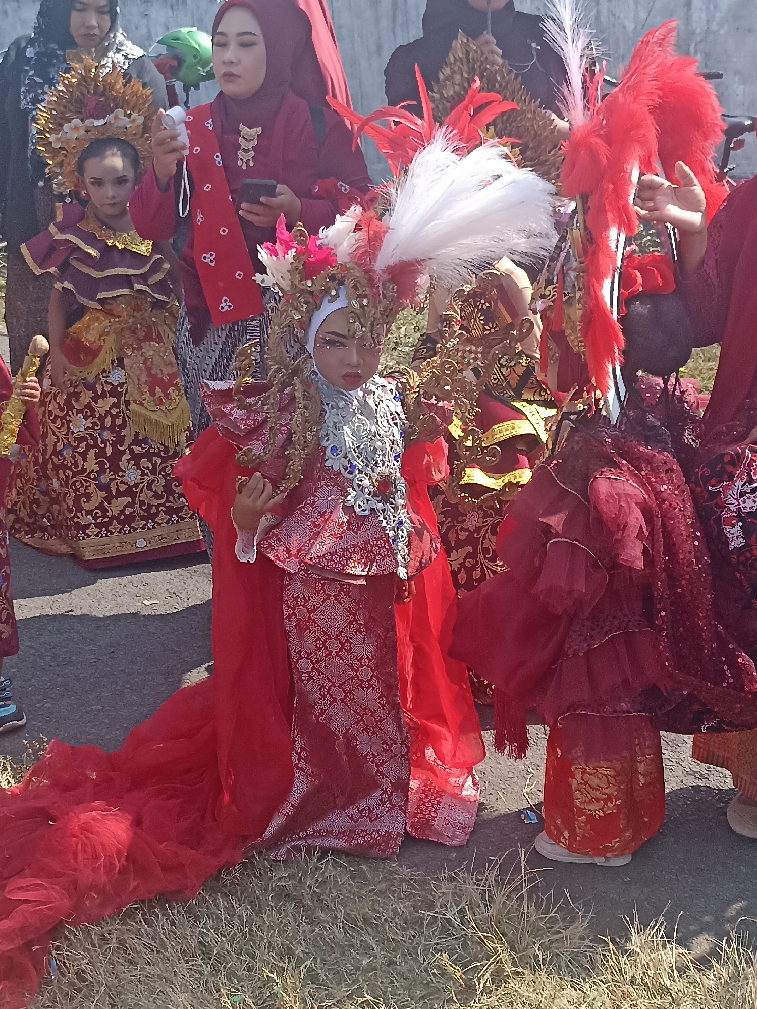 This is a medium shot, eye-level, outdoor, street-level photograph taken in Banyuwangi, Indonesia. The image features a group of people, predominantly women and children, dressed in elaborate, traditional red and gold ceremonial attire, suggesting a local festival or parade. In the foreground, a young child is the central focus, wearing a striking red costume with intricate gold patterns and a large, ornate headpiece adorned with white and red feathers. The child is positioned in the center of the frame, looking towards the camera with a serious expression. To the left of the central child, another young girl is visible, also in a detailed red and gold outfit and a golden headdress. Behind them, several adult women are present, some wearing headscarves and traditional clothing. One woman in the mid-ground is holding a mobile phone and a small white object. The attire is rich with detail, featuring layered fabrics, embroidery, and sparkling embellishments. The setting appears to be a paved street with some dry grass bordering the edge. The lighting suggests it is daytime, possibly late morning or afternoon. The overall atmosphere conveys a sense of cultural celebration and tradition.