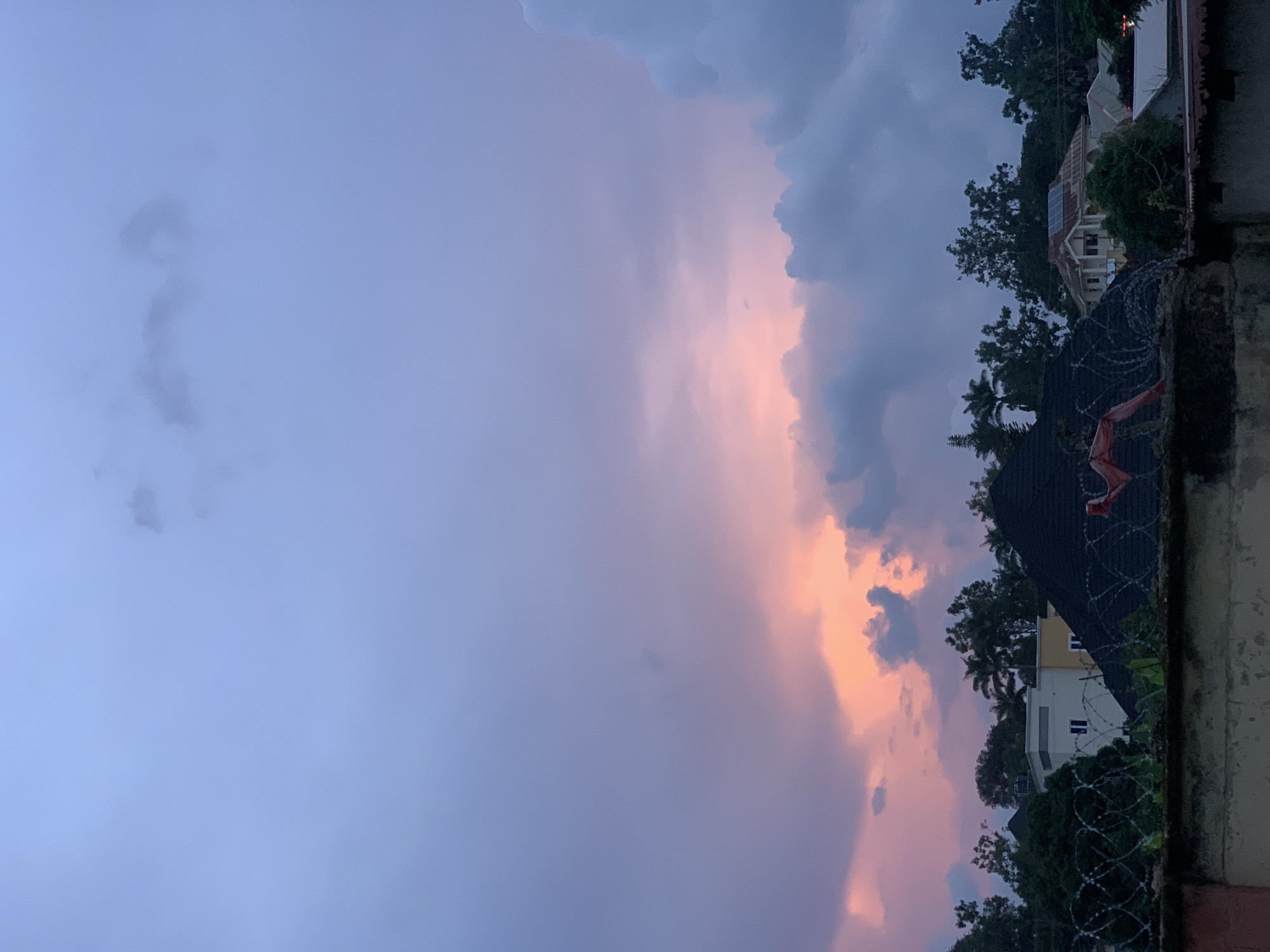 The image depicts a cloudy sky at dusk, with hues of purple, pink, and gray dominating the scene. A scattering of clouds forms a backdrop for the fading light, with a soft glow of pink and orange illuminating some of the cloud formations. Below the sky, the silhouettes of trees and rooftops are visible, suggesting an urban or semi-urban environment. The architecture of the buildings appears to be a mix of styles, with some featuring pitched roofs and others with flatter designs. A prominent dark, angular roof structure is in the foreground, and barbed wire fencing runs along the top of a wall, indicating a security measure. The overall impression is of a tranquil evening in a residential area, likely captured from an elevated position. The location is specified as the city of Jos, Nigeria.