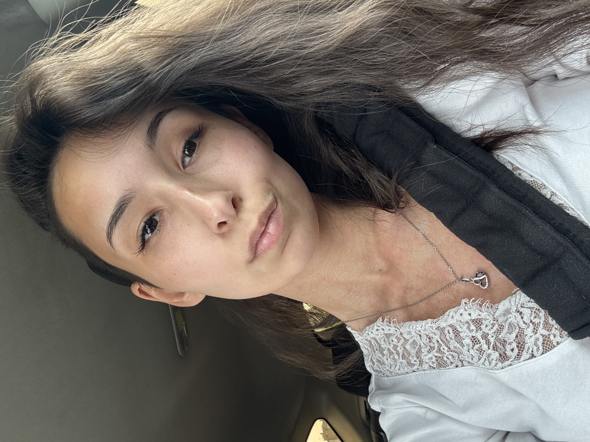 A young woman with dark hair and brown eyes is looking at the camera, her head tilted slightly to the side. She is wearing a white top with lace detailing and a silver necklace with a heart-shaped pendant. A black seatbelt is visible across her chest. The background appears to be the interior of a car. The lighting suggests it is daytime. The image was captured in Izhevsk, Russia.