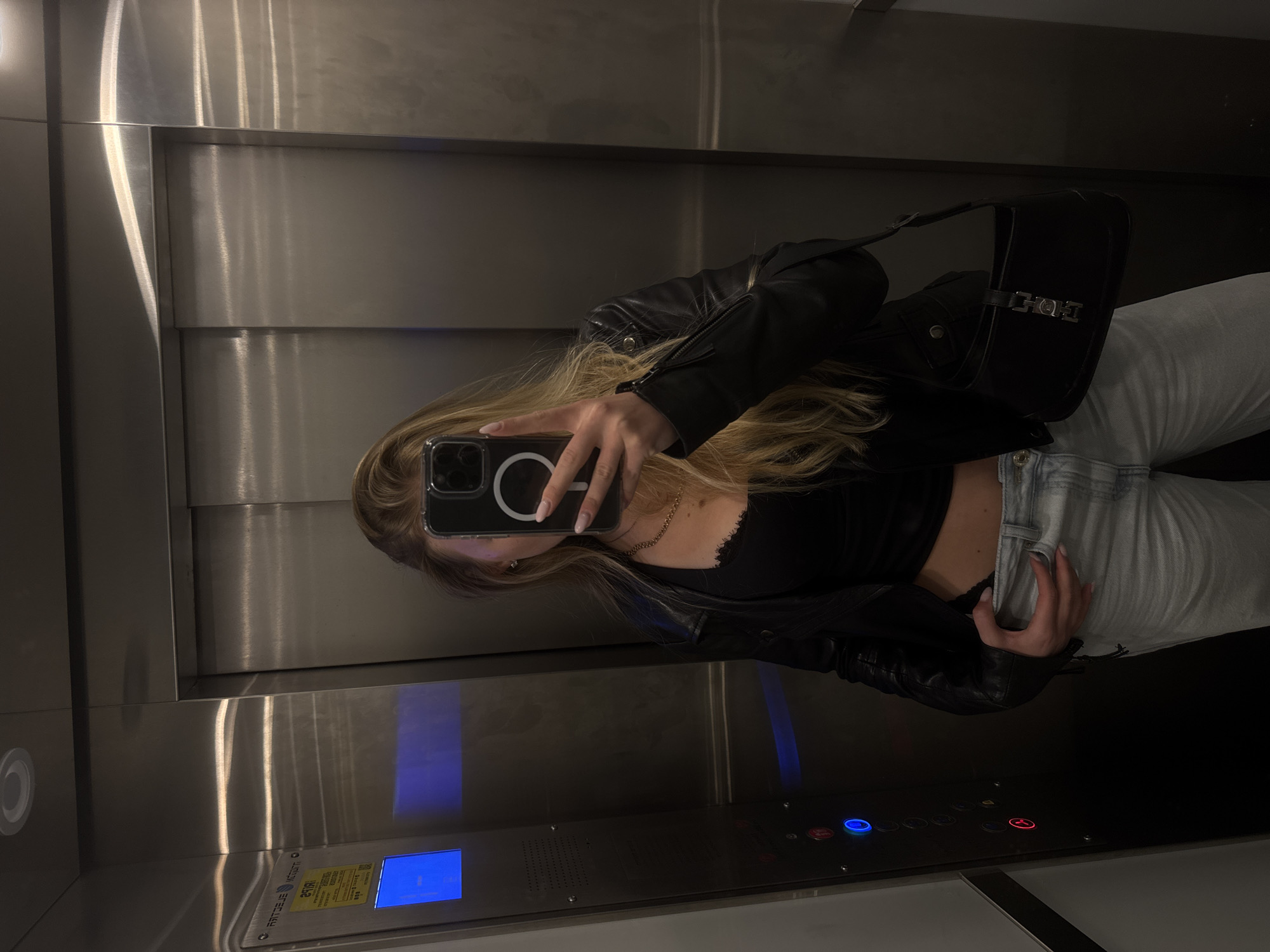 A young, light-skinned female takes a selfie in a metal elevator. She is wearing a black leather jacket over a black shirt, light-colored denim jeans, and has a black purse slung over her shoulder. Her hair is blonde and long. She is holding her phone, which appears to be a newer model iPhone, in front of her face to take the picture. The elevator is stainless steel with a control panel visible, featuring buttons with blue and red indicator lights. There is also a screen with text in Hebrew, indicating the elevator is likely in Tel Aviv, Israel.