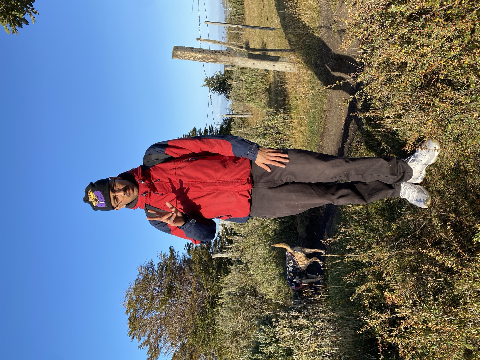 A man stands on a grassy path in Ushuaia, Argentina. He wears a red jacket, dark pants, white sneakers, glasses, and a black beanie with a purple and gold design. He makes a peace sign with his right hand. A wooden fence runs parallel to the path. Behind him, a dog in a patterned coat rests on a folded jacket. The sky is clear blue. The environment is a rural area with bushes and a distant mountain range.