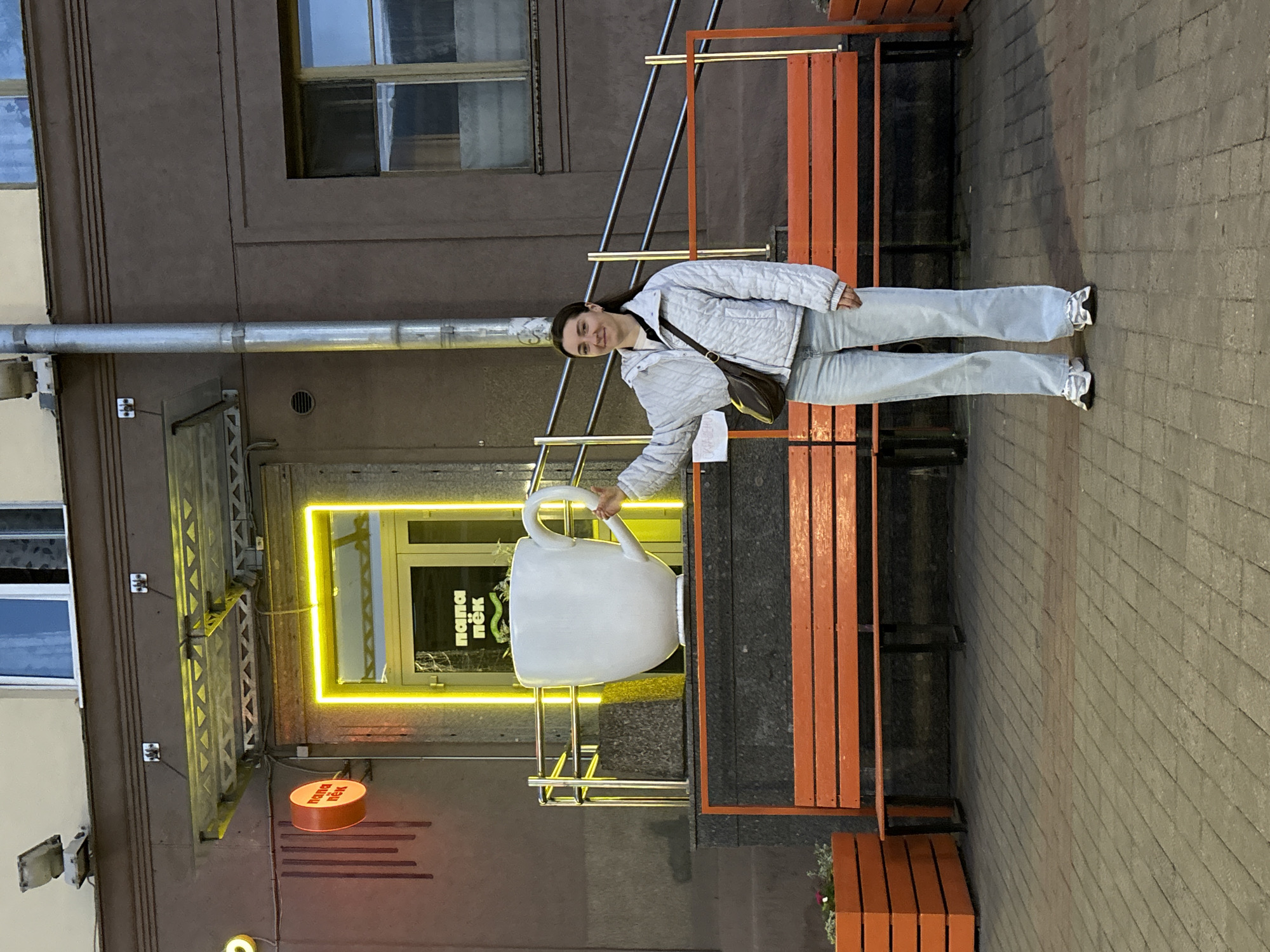 A young woman stands next to a large coffee cup sculpture in front of the "Папа Пёк" café in Minsk, Belarus. The café has a bright yellow neon sign. The woman is wearing a light-colored quilted jacket, jeans, and a dark brown shoulder bag. She is holding the handle of the giant coffee cup. The coffee cup sits next to an orange bench. The building is brown. The sidewalk is made of light gray bricks.