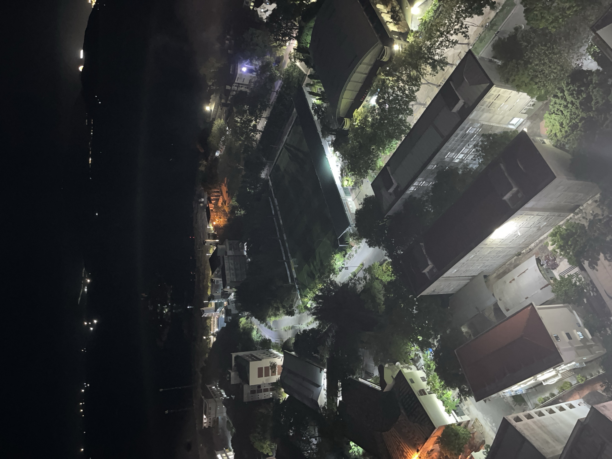 The image depicts an aerial view of a city at night. On the left is a dark body of water with scattered lights visible along the distant shoreline. Moving right, a cluster of buildings is seen illuminated by streetlights and building lights. The structures vary in size and style, with some featuring flat roofs and others peaked roofs. Trees are interspersed among the buildings, creating a mix of green and artificial light. Roads and pathways meander through the area, illuminated by streetlights. The overall environment is urban with a mixture of residential and commercial buildings.