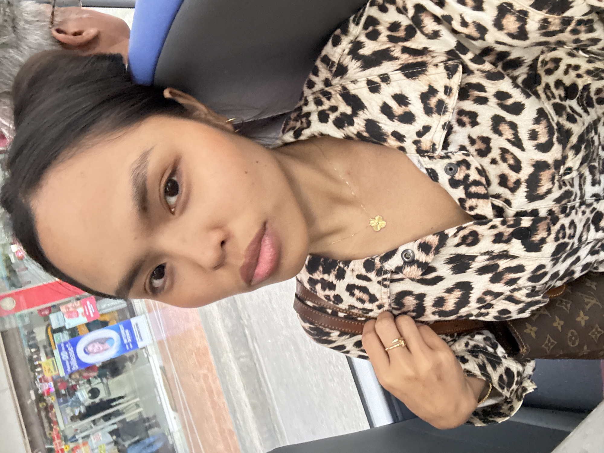 A young woman with dark hair pulled back is seated on a bus in Davao City, Philippines, facing forward and taking a selfie. She is wearing a leopard-print jacket, a gold clover pendant necklace, and a gold ring on her left hand. She carries a brown Louis Vuitton monogrammed purse over her left shoulder. Beside her, a man with gray hair and glasses is visible from the side, leaning his head against the seat. Through the bus window, a street scene with shops and pedestrians can be seen.