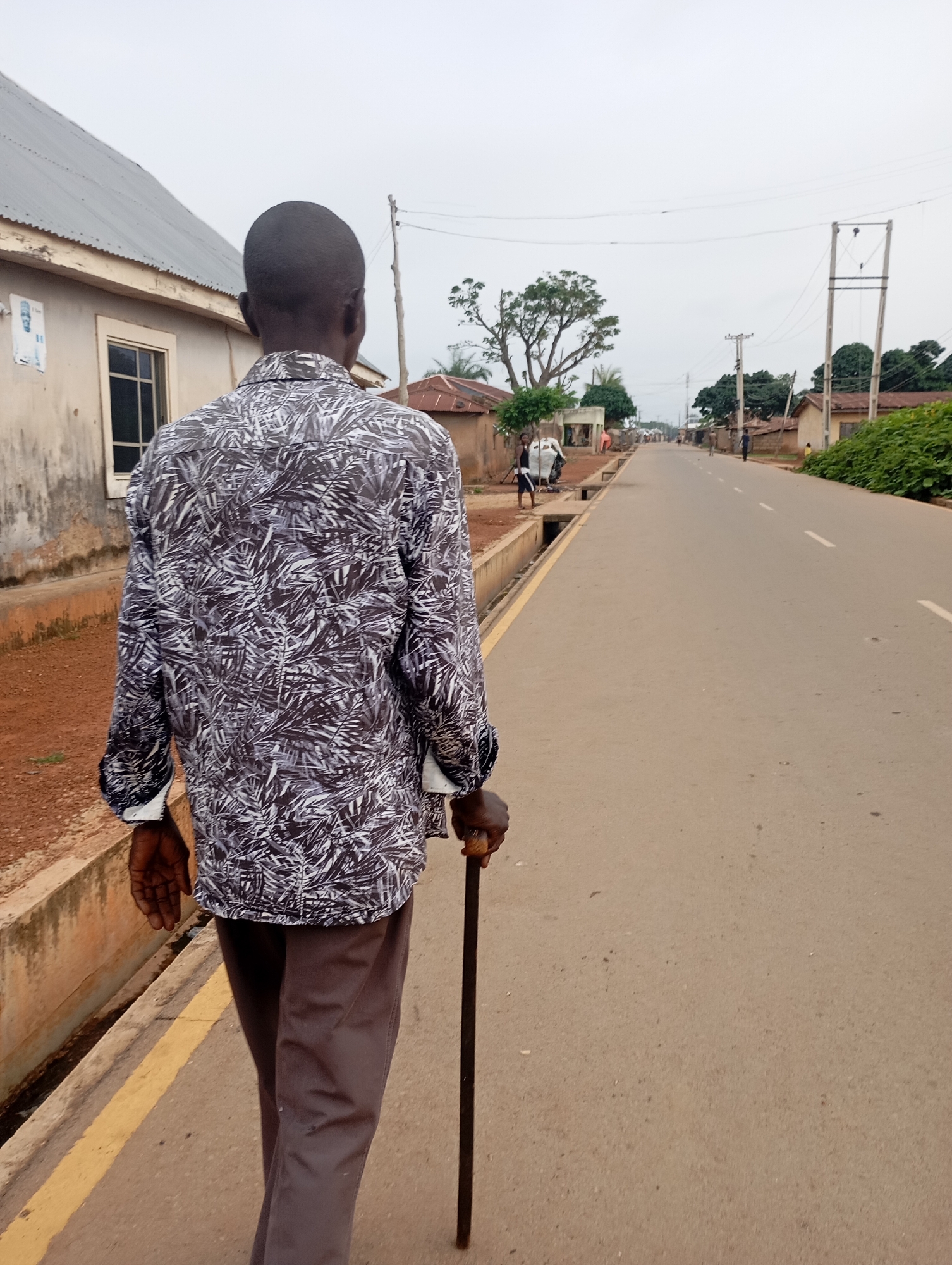 An over-the-shoulder, full-body shot depicts a Black man walking away from the camera on a paved road in Shendam, Nigeria. The man, who appears to be an elder, is wearing a patterned long-sleeved shirt and dark gray pants. He is holding a dark cane in his right hand for support. He is walking on the left side of the road, which has a yellow line marking the edge of the sidewalk. The road is lined with buildings and vegetation. In the background, there are power lines and poles, typical of the Nigerian infrastructure. The sky is overcast, suggesting a cloudy or hazy day. The overall scene captures a moment of everyday life in a Nigerian town.