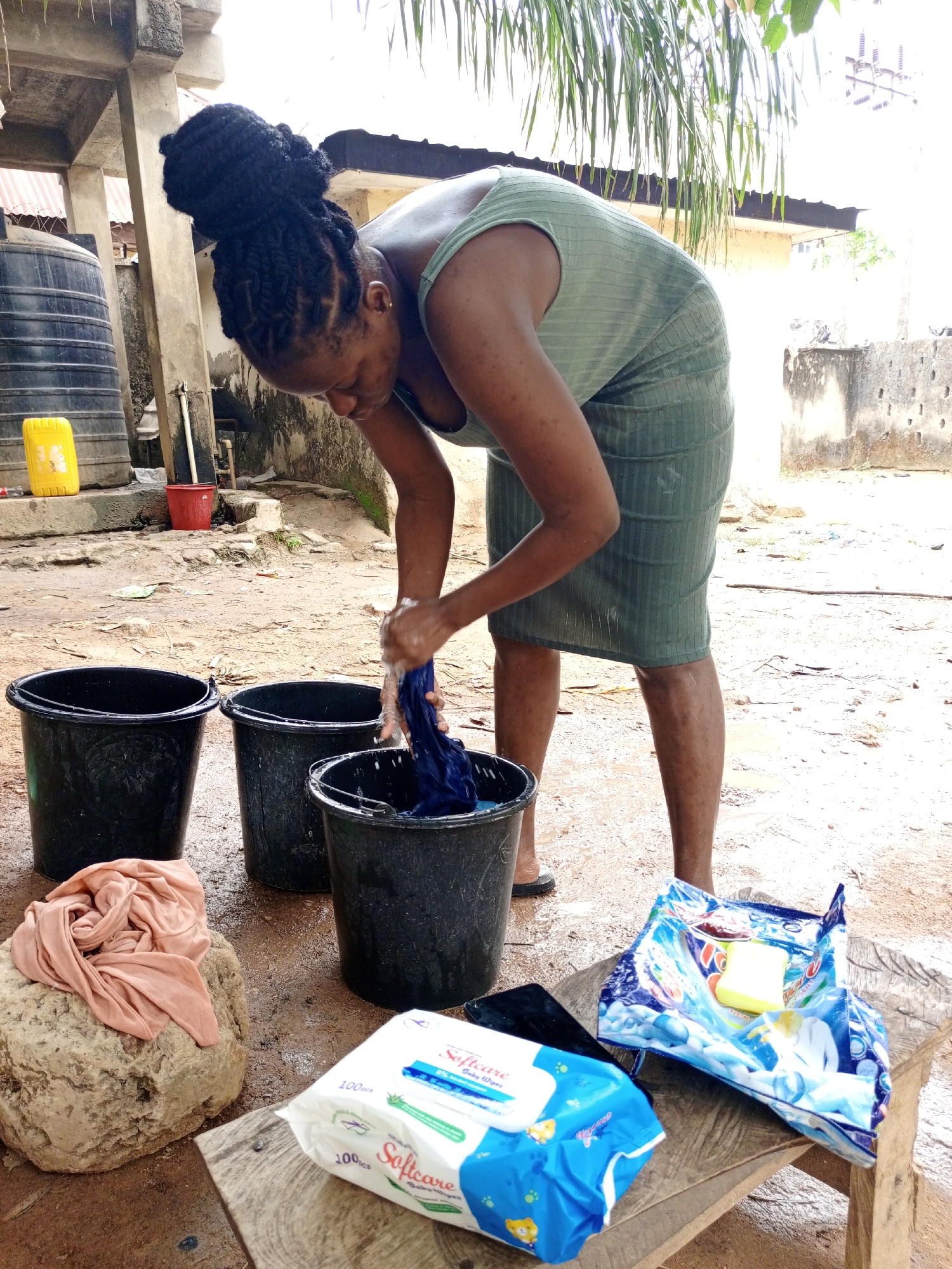 A woman is handwashing clothes outside in a compound in Keffi, Nigeria. She has dark hair braided and styled in a high bun. She is wearing a short, light green, bodycon dress. She is leaning over three black buckets, her hands immersed in one bucket with a piece of dark blue clothing. Three black buckets are placed near her. A light peach-colored fabric is placed on top of a large rock, and a pack of Softcare baby wipes, a bar of soap in a partly open bag, and a black smartphone are placed on a weathered wooden stool. A yellow jerry can is visible in the background near a large black water tank next to a building. The ground appears damp or wet with exposed dirt. Overhead, lush green foliage hangs into the frame. A concrete building is to the right.