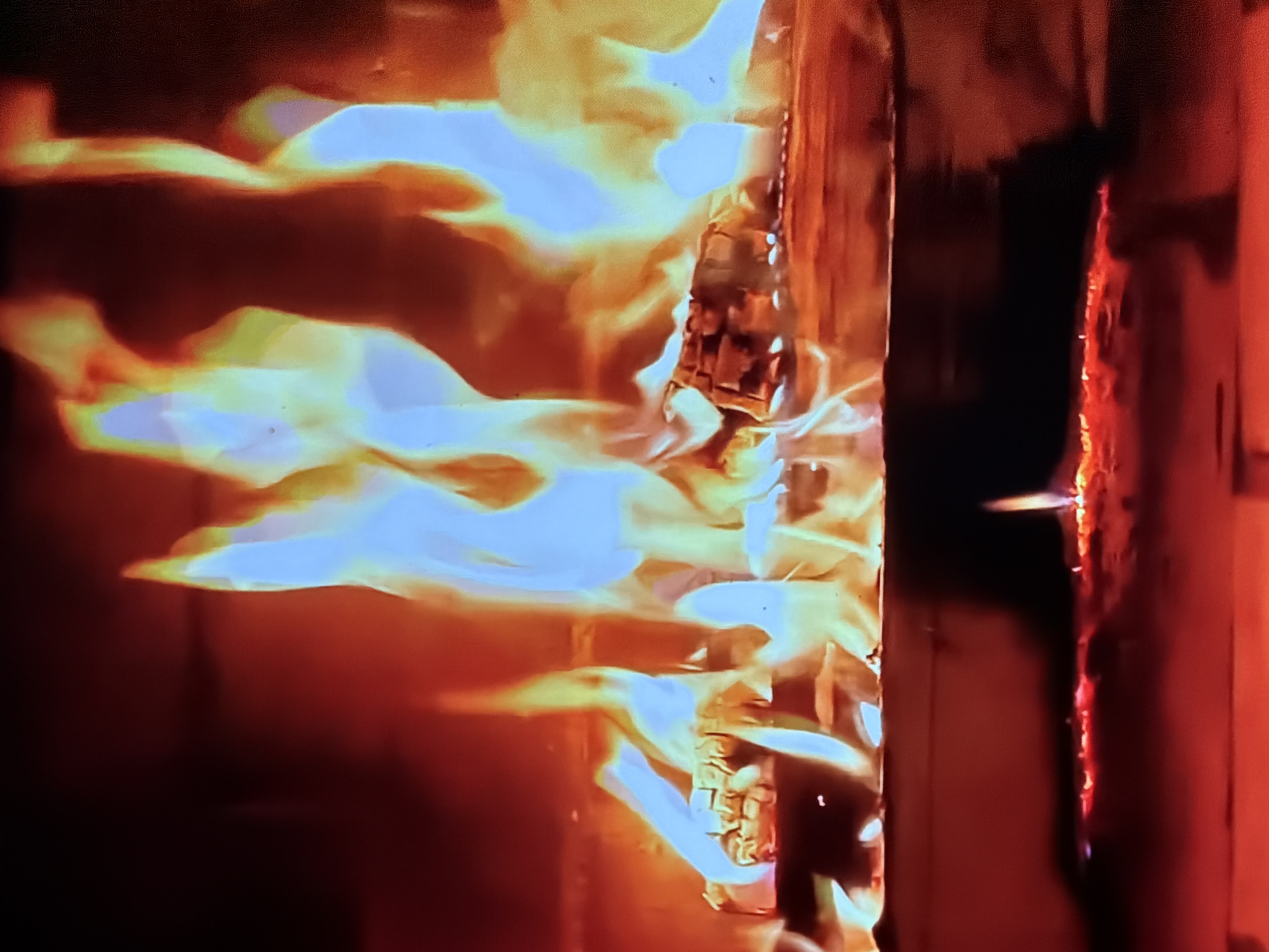 The image displays a contained fire, likely within a fireplace or fire pit. Dynamic, irregular flames dominate the upper two-thirds of the frame, exhibiting distinct shades of bright blue, yellow, and orange, indicating varying temperatures of combustion. Multiple wooden logs, some actively burning and others charred, form the primary fuel source, visible in the mid-ground. Beneath the logs, a glowing bed of intense red and orange embers extends horizontally across the lower portion of the image. A small, distinct, upward-reaching flame emerges from this ember bed on the lower-right side. The immediate surroundings of the fire are dark, illuminated only by the fire's radiant glow, which casts a reddish hue on the internal surfaces of the containment structure. The location of this scene is in the city of Maidstone, United Kingdom.