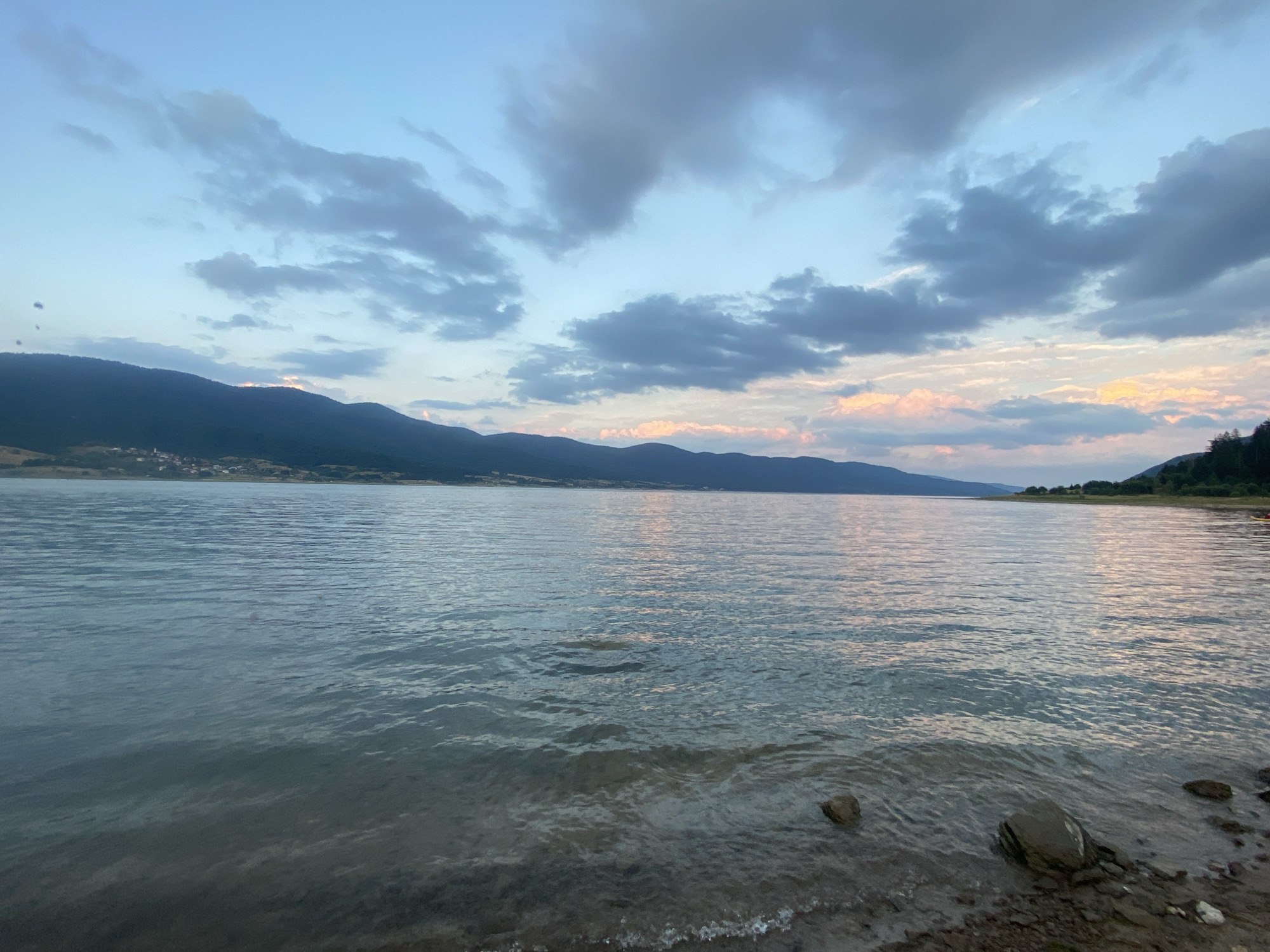 The image depicts a serene natural landscape by a body of water, likely a reservoir or lake, with mountains in the background. The time of day appears to be dusk or dawn, with soft, diffused light illuminating the sky and reflecting on the water's surface. The sky is a gradient of blue, with scattered clouds tinged with pink and orange hues, suggesting the presence of the setting or rising sun.
In the mid-ground, a range of rolling hills or mountains forms a dark silhouette against the sky. A small village or settlement with scattered lights is visible nestled on the slopes of the mountains on the left side of the image. The water in the foreground is calm, with gentle ripples and reflections of the sky and distant landscape. The shoreline is rocky and appears to be covered with small pebbles and stones. The overall atmosphere is tranquil and picturesque.
The location is identified as Sarnitsa, Bulgaria, a town situated on the shores of the Dospat Reservoir, a large artificial lake in the Rhodope Mountains. The scene captured is characteristic of the natural beauty found in this region, known for its reservoirs, forests, and mountain vistas.