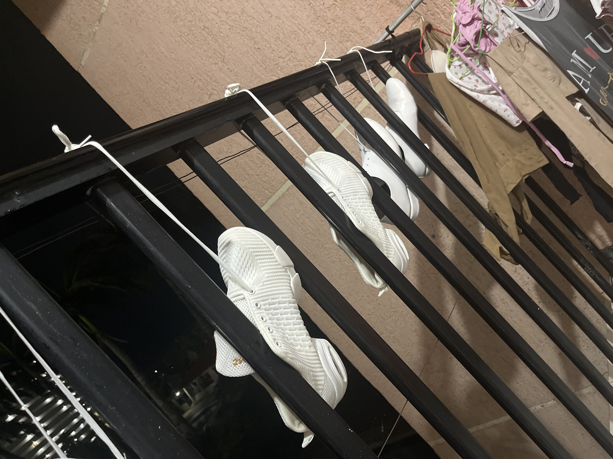 The image captures a scene of shoes and clothes air-drying on a black metal railing, likely on a balcony or an outdoor drying rack, at night in Porlamar, Venezuela.
In the foreground, several pairs of shoes are hung. Two pairs of white athletic shoes, or sneakers, are prominently displayed, with their white laces used to loop them over the horizontal bars of the railing. The sneakers feature a textured, woven upper material. A small gold logo or text, possibly "220" or a brand name, is visible on the side of one sneaker. Further along the railing to the right, a pair of white ballet flats or similar dress shoes is also hung.
Beyond the shoes, various clothing items are draped over or hanging from the railing. These include a pair of light brown or khaki pants, and several pieces of fabric in pink and a patterned white with pink, possibly children's clothes or undergarments, some hanging from pink hangers. A grey fabric bag or basket, partially visible on the right, also holds clothing, with the word "LAUNDRY" partially discernible on its side.
The railing itself is composed of black horizontal bars supported by vertical posts, suggesting a robust structure. The background indicates it is nighttime, with a dark sky and blurred distant lights from what appear to be other buildings or streetlights, hinting at an urban or suburban environment. The ground or wall directly behind the railing appears to be a light brown or beige color, possibly tiled. The overall impression is one of domestic activity taking place outdoors after dark.
