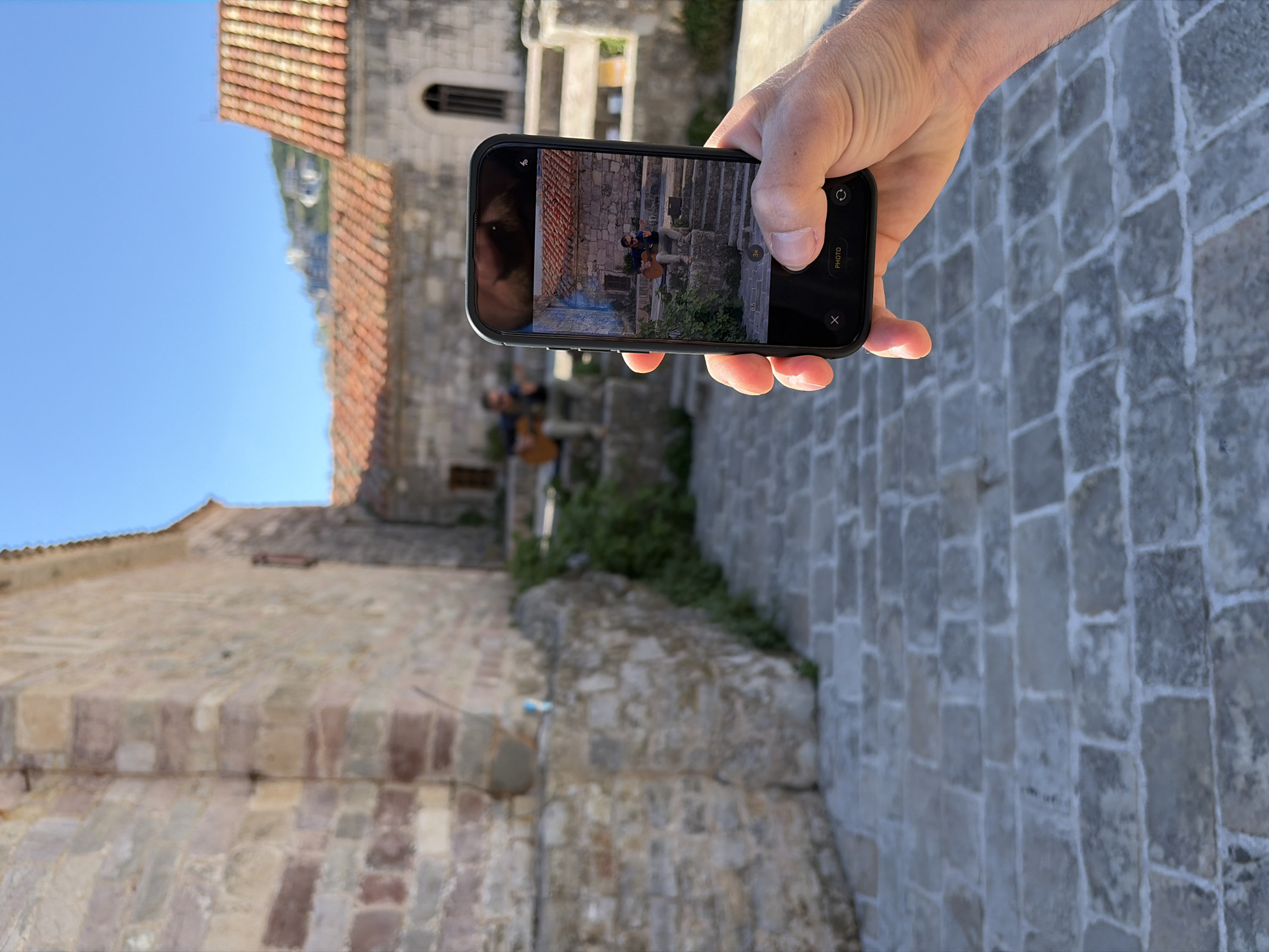 Here's a detailed description of the image:
**Setting:**
The image appears to be taken in a historic urban environment, likely in the city of Budva, Montenegro, based on the architectural style. Stone buildings with red tile roofs are visible. The environment appears to be an old town or a historic district.
**Objects:**
* **Phone:** A person is holding a smartphone with its screen displaying an image.
* **Stone Structures:** The primary architecture in the scene consists of buildings and a walkway constructed from stone blocks.
* **Vegetation:** Some green vegetation is growing on or near the stone structures.
**Subjects and Actions:**
* **Person Taking a Photo:** A hand is holding the phone. The person is taking a photo of another person sitting on stairs with a guitar.
* **Person in the Phone's Screen:** On the screen of the phone, there is an image of a person sitting on stone stairs playing a guitar.
**Interaction:**
The person is capturing an image of the person playing music.
**Other details:**
*The person in the phone is taking a photo of a musician playing a guitar on the stairs and this musician is the same one as in the phone, the person taking the picture is recording a loop.*