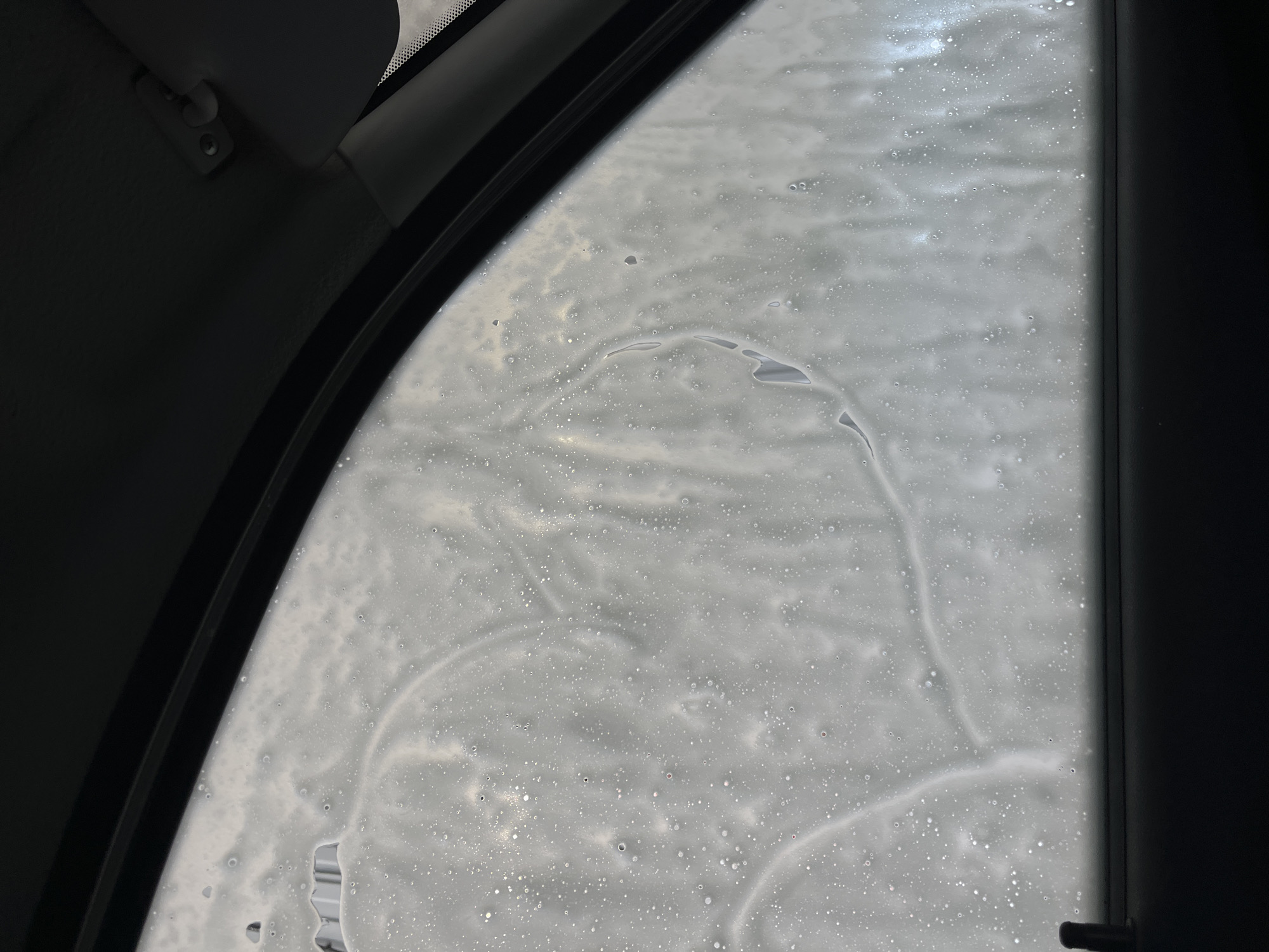 The image shows a view from inside a car, looking out through the side window. The window is covered in soapy water, likely from a car wash. The car's interior is visible along the edges of the window frame, including the sun visor attachment. No people or specific objects beyond the car and soap are identifiable. The location within Chaikovsky, Russia cannot be determined from the given information.