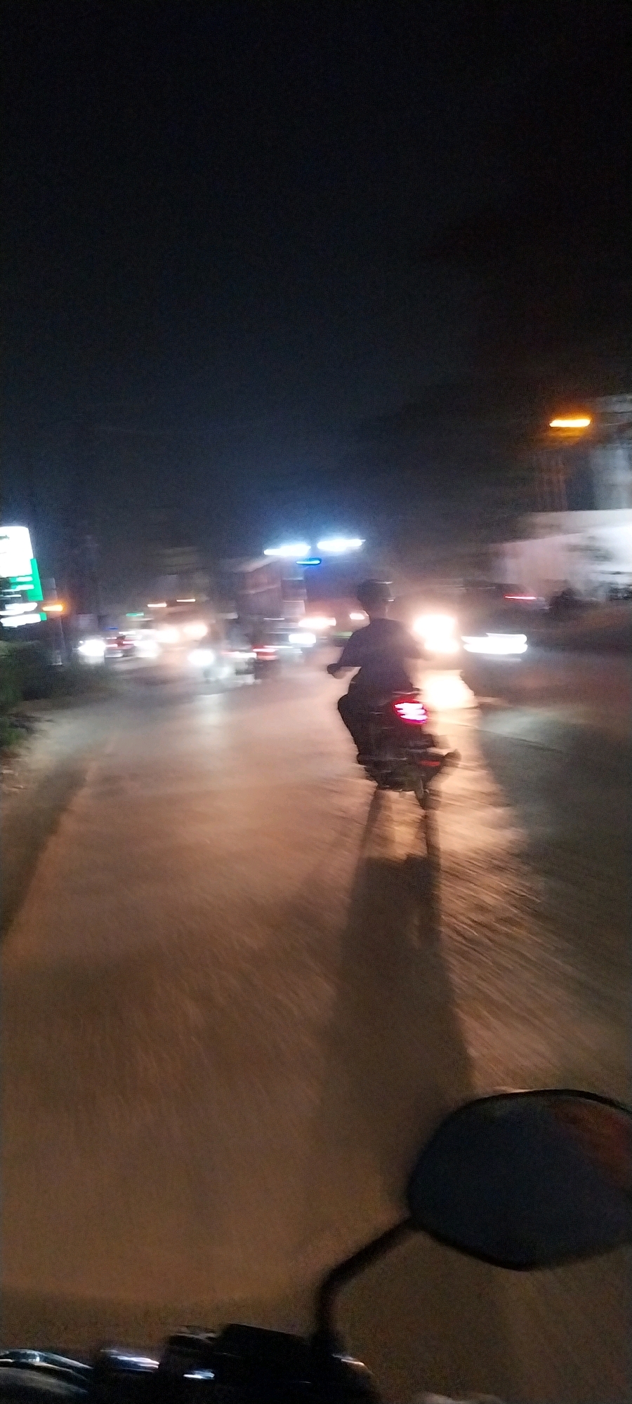 An over-the-shoulder shot from a motorcycle at night shows a street scene in Kelapa Dua, Indonesia. The road is wet and reflects the lights of passing vehicles. The main subject is another motorcycle moving away from the camera, with a rider visible. The rider appears to be wearing dark clothing. In the background, there are blurry lights from buildings, streetlights, and other traffic. The overall impression is a fast-moving, nighttime scene with limited visibility due to the darkness and motion blur.