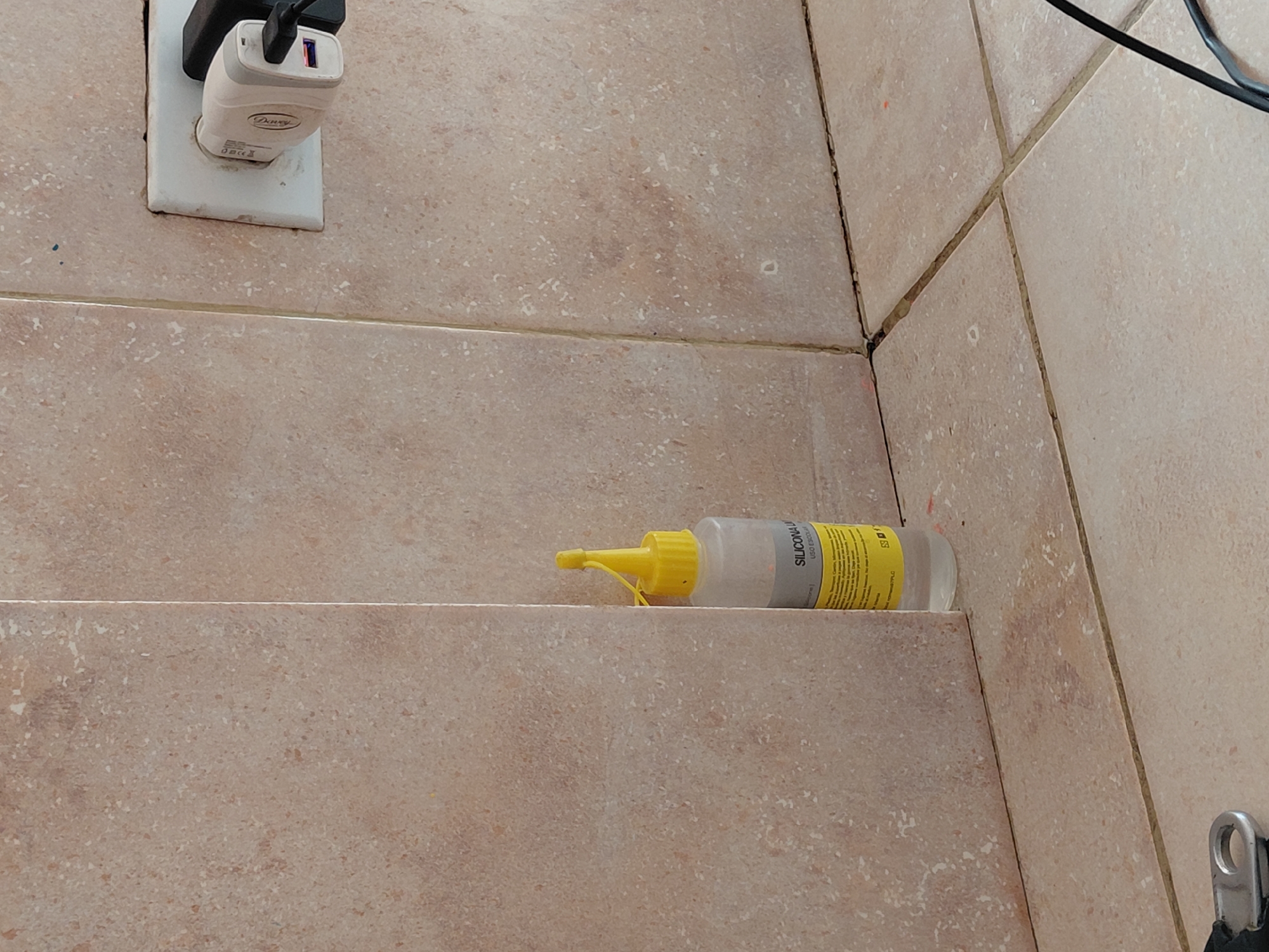 An eye-level shot captures a section of a tiled wall and floor, likely indoors in Santa Ana, Venezuela. The tiles are a light beige or terracotta color with a speckled, worn appearance, and visible grout lines, some of which show discoloration.
In the upper left, a white electrical outlet is mounted on the tiled surface. A white USB wall charger, faintly branded "Daway," is plugged into the outlet. It features two USB ports, one with a black interior and the other with a pink/purple interior, and a black cable is connected to one of the ports, extending upwards out of frame. The charger also displays regulatory symbols such as CE and a crossed-out trash can.
Horizontally positioned on a tiled ledge or step in the center-right is a clear plastic bottle with a bright yellow conical cap and nozzle. The bottle contains a clear liquid. A white label with yellow text is wrapped around it, clearly showing the words "SILICONA LIQUIDA" (liquid silicone) and "USO ESCOLAR" (school use), indicating it is likely a craft adhesive or sealant.
In the bottom right corner, a silver-colored metal loop or hook is partially visible, attached to an unseen object. The overall scene suggests a utility area or a corner of a room where repairs or crafting might take place, given the presence of the silicone bottle and the electrical charging setup. The lighting appears to be ambient indoor light.