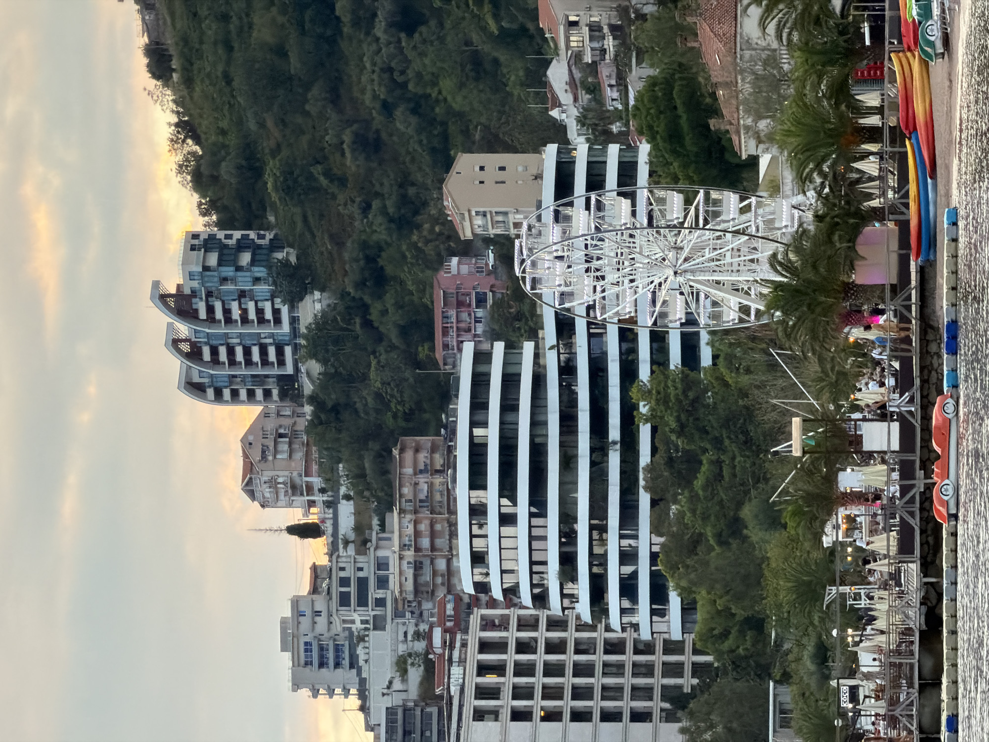 This is a telephoto shot of the Budva Riviera in Montenegro, captured during the golden hour. The scene depicts a vibrant urban landscape against a backdrop of lush green hills. Dominating the foreground is a white Ferris wheel, a popular tourist attraction, situated on the waterfront. Adjacent to it, a beach is lined with colorful kayaks and pedal boats, hinting at recreational activities. Beyond the waterfront, a series of modern buildings, characterized by their white facades and horizontal architectural lines, rise up the hillside. Some of these buildings feature large, reflective windows, suggesting they are hotels or residential apartments. In the background, older, more traditional houses with terracotta roofs are interspersed with the modern architecture and dense vegetation. The sky is a soft gradient of orange and yellow, illuminated by the setting sun, creating a warm and inviting ambiance. A few people are visible on the beach and along the waterfront promenade, engaged in casual strolling. The overall impression is one of a bustling seaside resort town transitioning into evening.