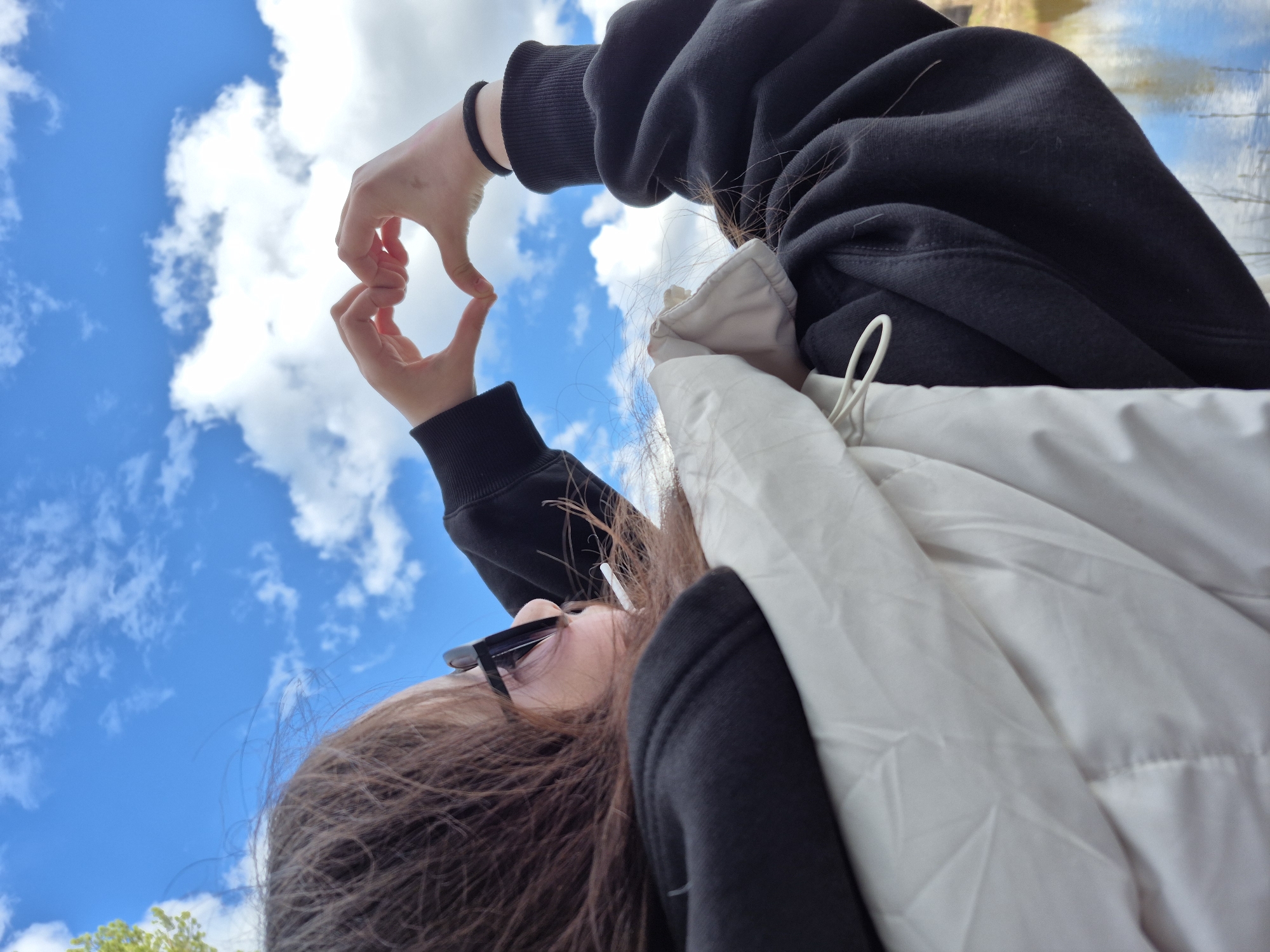 A person, seen from a low-angle view, is holding their hands up to form a heart shape against a backdrop of a blue sky with scattered white clouds. The person is wearing a black sweatshirt and a white, possibly puffy, vest or jacket. They also have sunglasses on. The location is Chaikovsky, Russia. The focus is on the gesture of making a heart shape with the hands, symbolizing love or affection towards the sky.