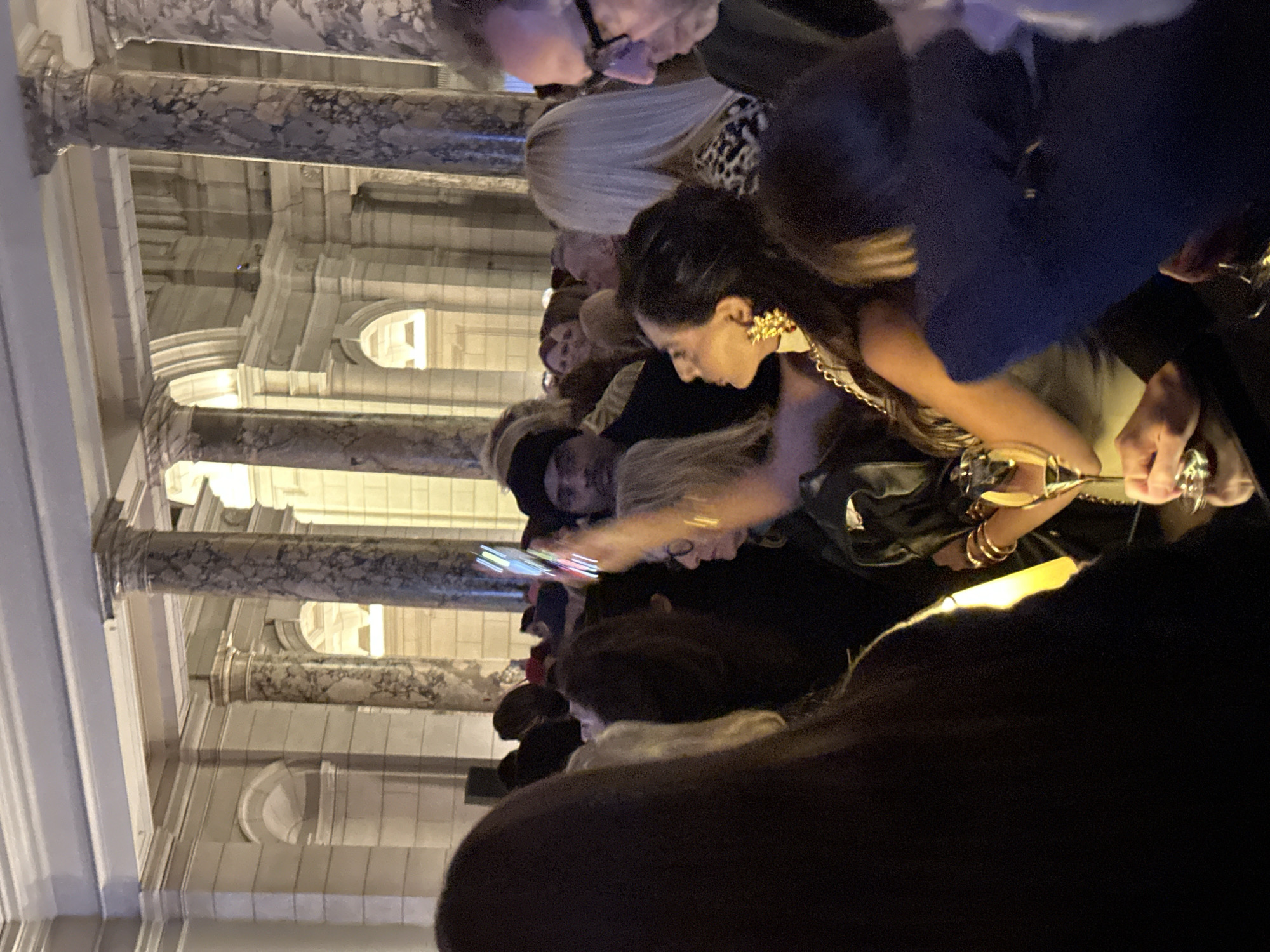 The scene depicts a crowded indoor social event within a grand architectural setting, identified as the Victoria and Albert Museum (V&A) in South Kensington, London, United Kingdom. The background features multiple large, classical columns, some appearing to be veined marble, and arched alcoves, illuminated by warm, ambient light. In the mid-ground, a person with dark skin and a dark head covering looks directly forward. To their immediate right, a woman with long dark hair is visible in profile, wearing a large, ornate gold earring and holding a stemmed glass containing a light-colored beverage. Her left wrist displays multiple gold-toned bracelets. Above her, a mobile phone is held aloft, its screen emitting bright, multi-colored vertical light streaks. To the far right, a man with glasses looks downwards. Multiple other individuals are partially visible in the dense crowd, indicating a social gathering or reception.