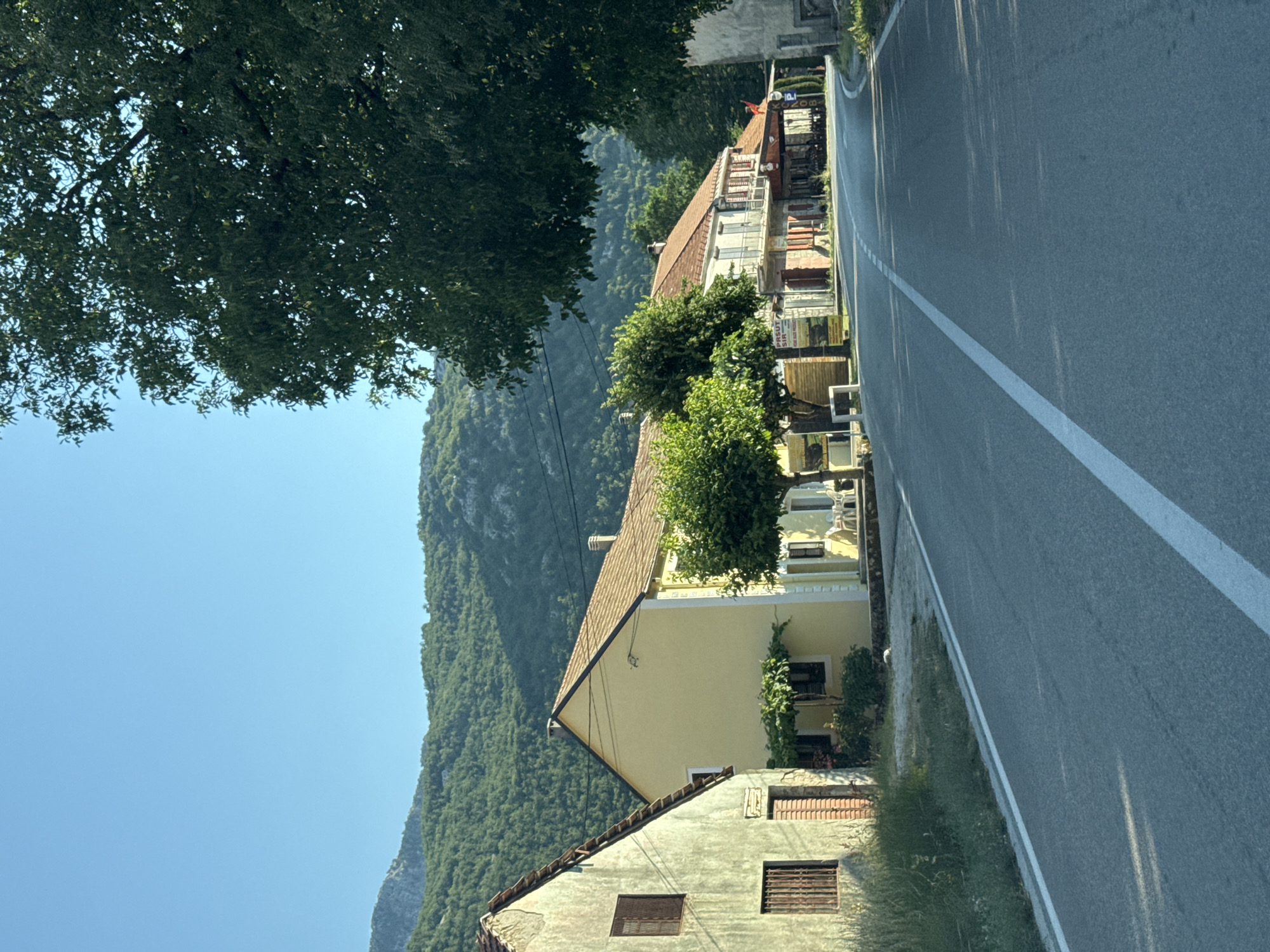 This is a long shot, eye-level, outdoor, daylight, vertical, eye-level, medium shot of a road leading through a village. In the foreground, a paved road with white lines runs towards the center of the frame. The road curves to the right as it recedes into the distance. On the left side of the road, there are several buildings. The closest building is a light yellow house with a brown roof. Next to it is a pale green building with brown window frames. Further down the road, on the right side, are more buildings with awnings and signs, some of which appear to be shops or restaurants. The buildings are a mix of colors, including yellow, white, and brown. Green trees are visible in front of some of the buildings and also overhanging the road in the upper right portion of the image. In the background, a lush green, tree-covered mountain range rises under a clear blue sky. The scene appears to be in a rural area, possibly a village in Montenegro. The time of day appears to be morning or late afternoon, given the angle of the sunlight. There are no people visible in the image.