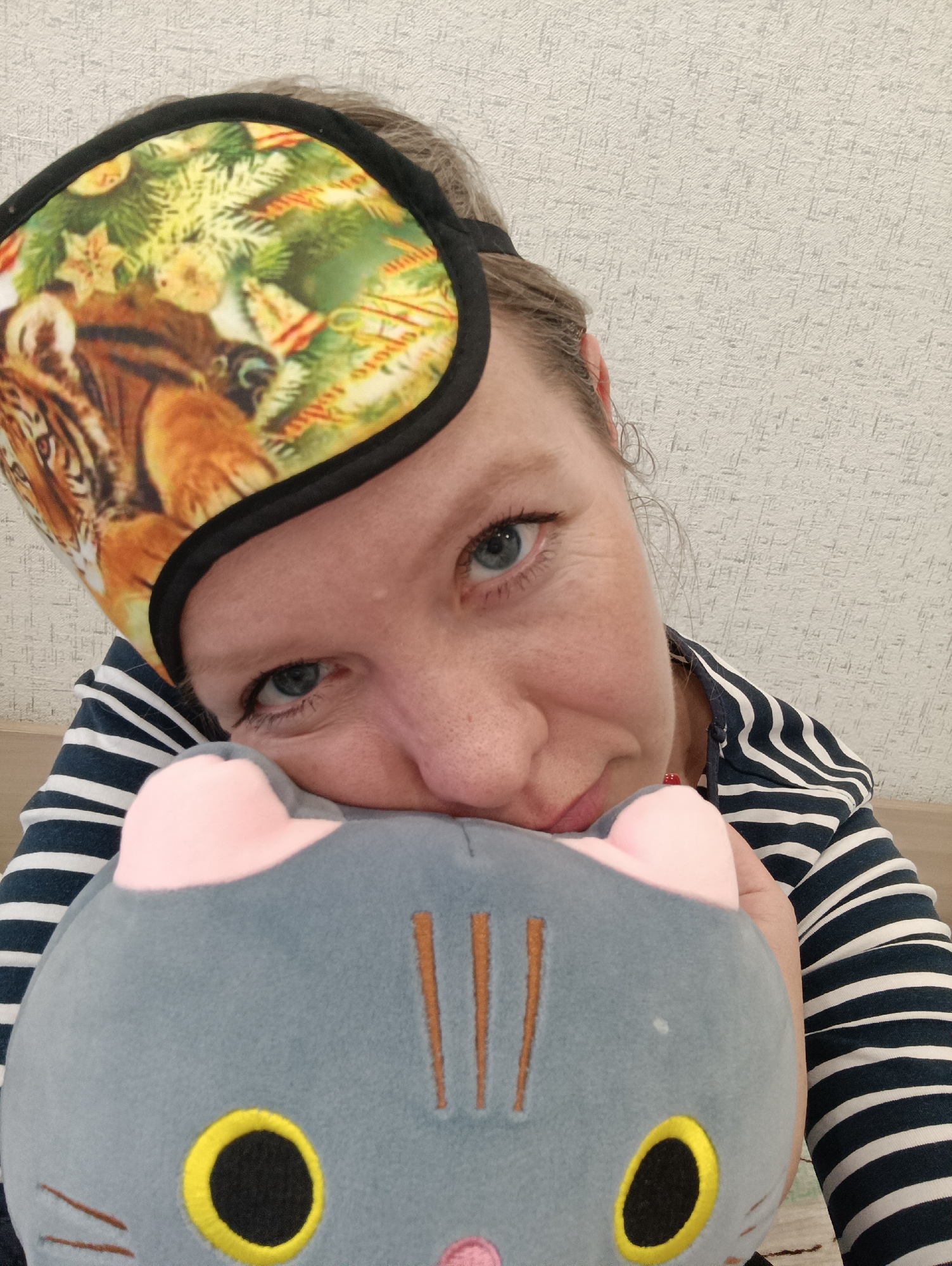 A close-up, eye-level portrait shows a woman indoors, likely in Chaikovsky, Russia, looking directly at the camera with a gentle expression. Her blue eyes are prominent, and her light-colored hair is visible at the sides and top.
She wears a colorful sleep mask pushed up onto her forehead. The mask features a bold design incorporating a tiger's face on the left side, blended with festive elements like green Christmas tree branches, golden stars, red ornaments, and party hats. The mask has a black trim.
The woman is dressed in a dark blue top or hoodie with white horizontal stripes. She is holding a grey cat plush toy close to her, resting her chin softly on its head. The plush toy has pink ears, distinct yellow-rimmed black oval eyes, and brown embroidered stripes or whiskers.
The background is a light-colored wall with a subtle, textured pattern, indicating an indoor setting. The overall mood is casual and comfortable, suggesting a moment of relaxation. There is no visible text in the image.
