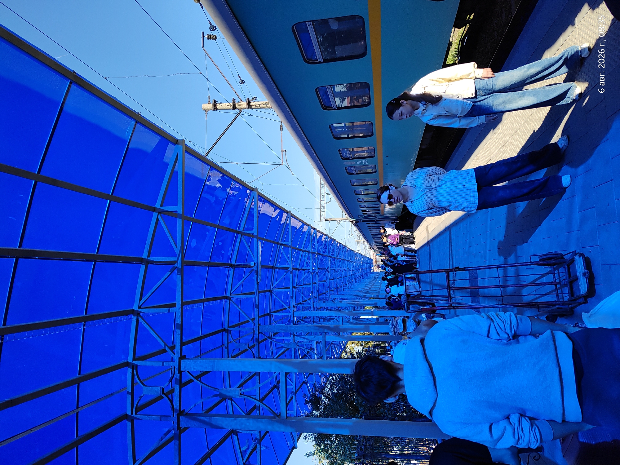An outdoor, eye-level, medium shot captures a sunny day at a train station in Astana, Kazakhstan. A vibrant blue canopy with metal supports extends over the platform, casting a cool blue hue on the scene. To the right, a teal train with a yellow stripe runs along the tracks, its windows reflecting the surroundings. Several people are visible on the platform, some standing, others walking. In the foreground, a person wearing a grey hoodie is seen from behind. Two young women, one in a striped shirt and dark jeans, the other in a light jacket and denim jeans, stand near the train. A luggage cart with red metal bars is positioned on the platform. The ground is paved with patterned tiles. The sky is clear and bright blue. A timestamp in the lower right corner reads "6 авг. 2026 г., 08:35".
