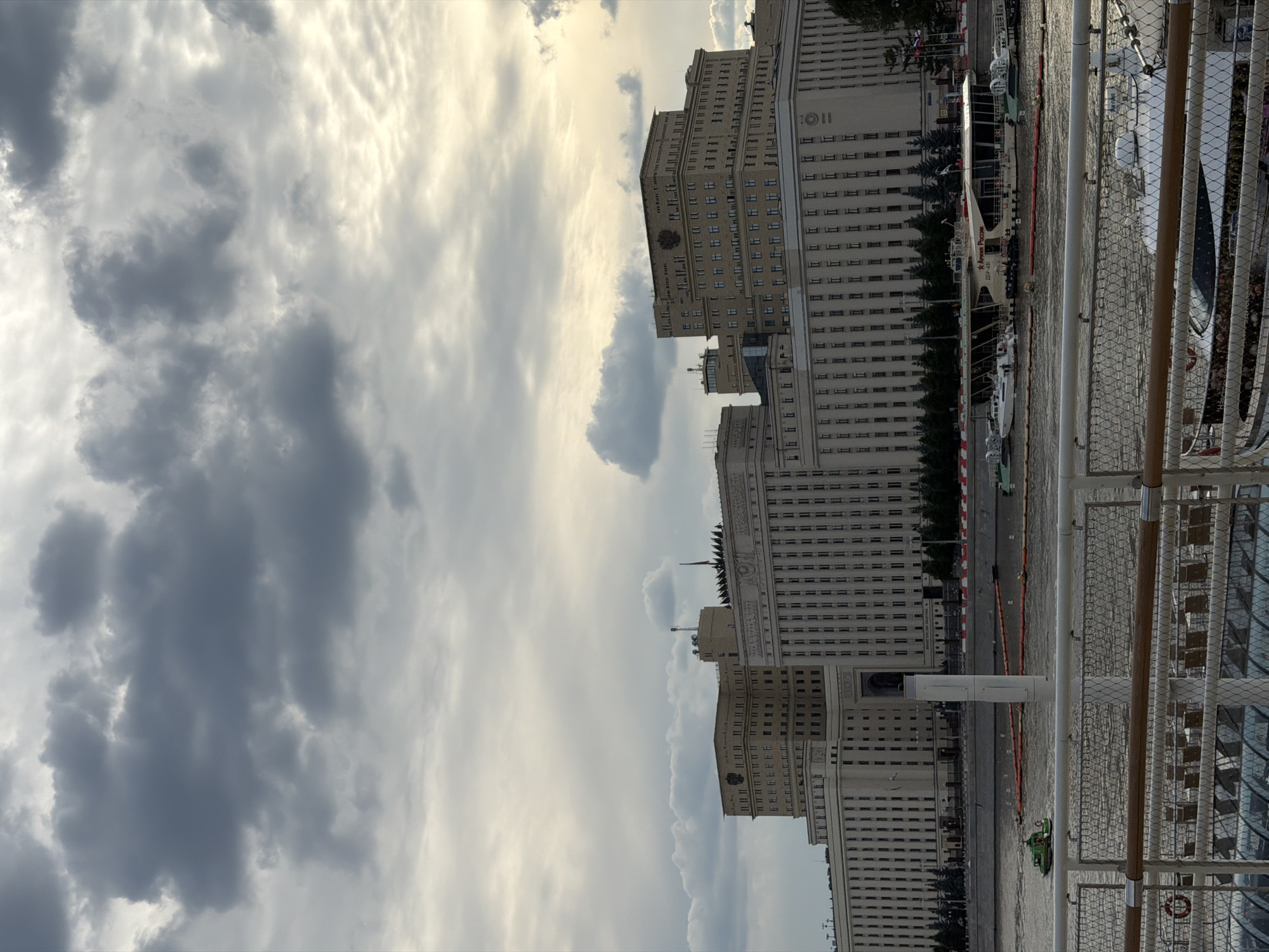 The image captures a waterfront view of the Ministry of Defense building in Moscow, Russia. The massive, Stalinist-era complex dominates the skyline, characterized by its numerous windows and imposing architecture. In the foreground, a river separates the viewer from the building, with several boats visible on the water. A small green boat is seen moving against the current, while larger vessels, including what appears to be a restaurant boat with "АРМИЯ РОССИИ" (Army of Russia) emblazoned on its side, are docked or moving slowly. The sky above is filled with dramatic, dark clouds, suggesting an overcast or approaching storm, with a sliver of sunlight breaking through on the right side of the frame, casting a warm glow on the clouds. The overall atmosphere is one of grandeur and historical significance, set against a dynamic natural backdrop.