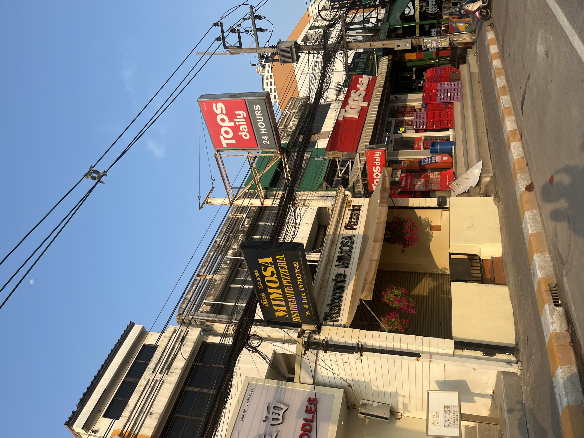 The image depicts a commercial street scene in Pattaya City, Thailand, under a clear sky. Several storefronts are visible, with "Tops daily" convenience stores being prominent, indicated by red and white signage with "24 HOURS" noted on one sign. A "MIMOSA RISTORANTE PIZZERIA" sign is also seen, advertising Italian cuisine, along with contact information. Below Mimosa is another storefront with a sign in Thai and English that reads "Noodles." Red and purple plastic crates are stacked near the entrance of one of the Tops Daily stores, and an electrical utility pole is adorned with many wires. The architecture is a mix of low-rise commercial buildings, with an older aesthetic visible on some of the facades. The street is paved, with a curb painted in alternating white and yellow stripes. A partial view of the moon is visible in the upper left corner of the sky.