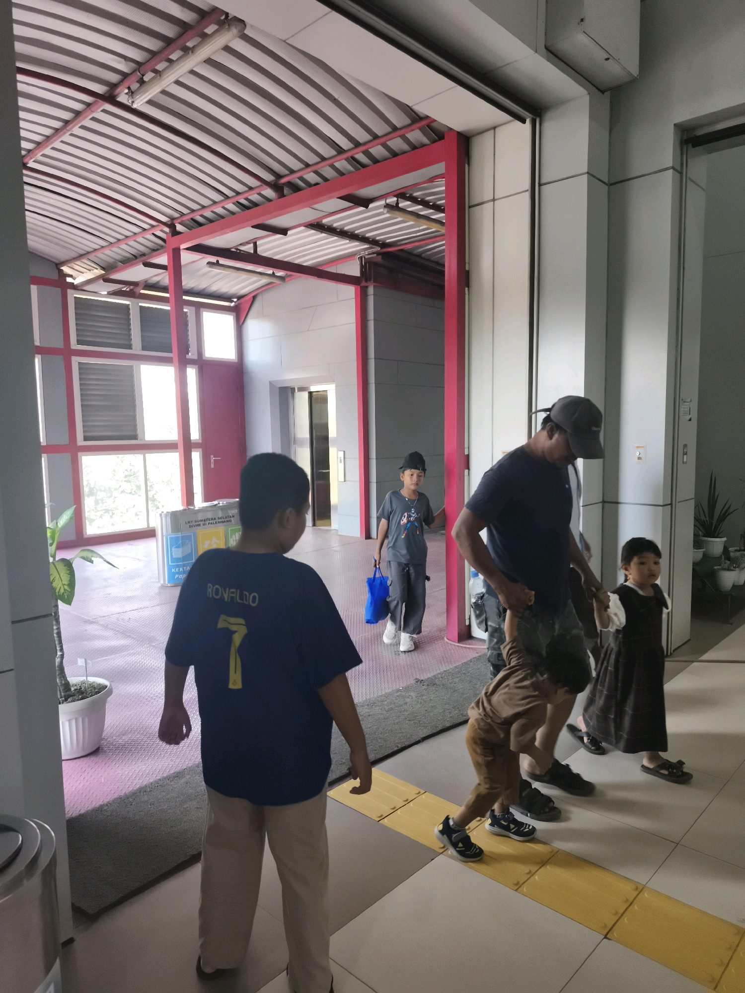 A group of people, including adults and children, are walking down a hallway in a building. The hallway has a corrugated metal ceiling with red supports. There are windows and doors visible in the background, as well as a plant in a white pot. A trash can is also visible on the left side of the image. The floor is tiled with a yellow tactile warning strip. The group appears to be heading towards an elevator on the right side of the image. The scene is consistent with an indoor public space such as a shopping mall or a public building in Kelurahan 16 Ilir, Kec. Ilir Timur I, Indonesia.