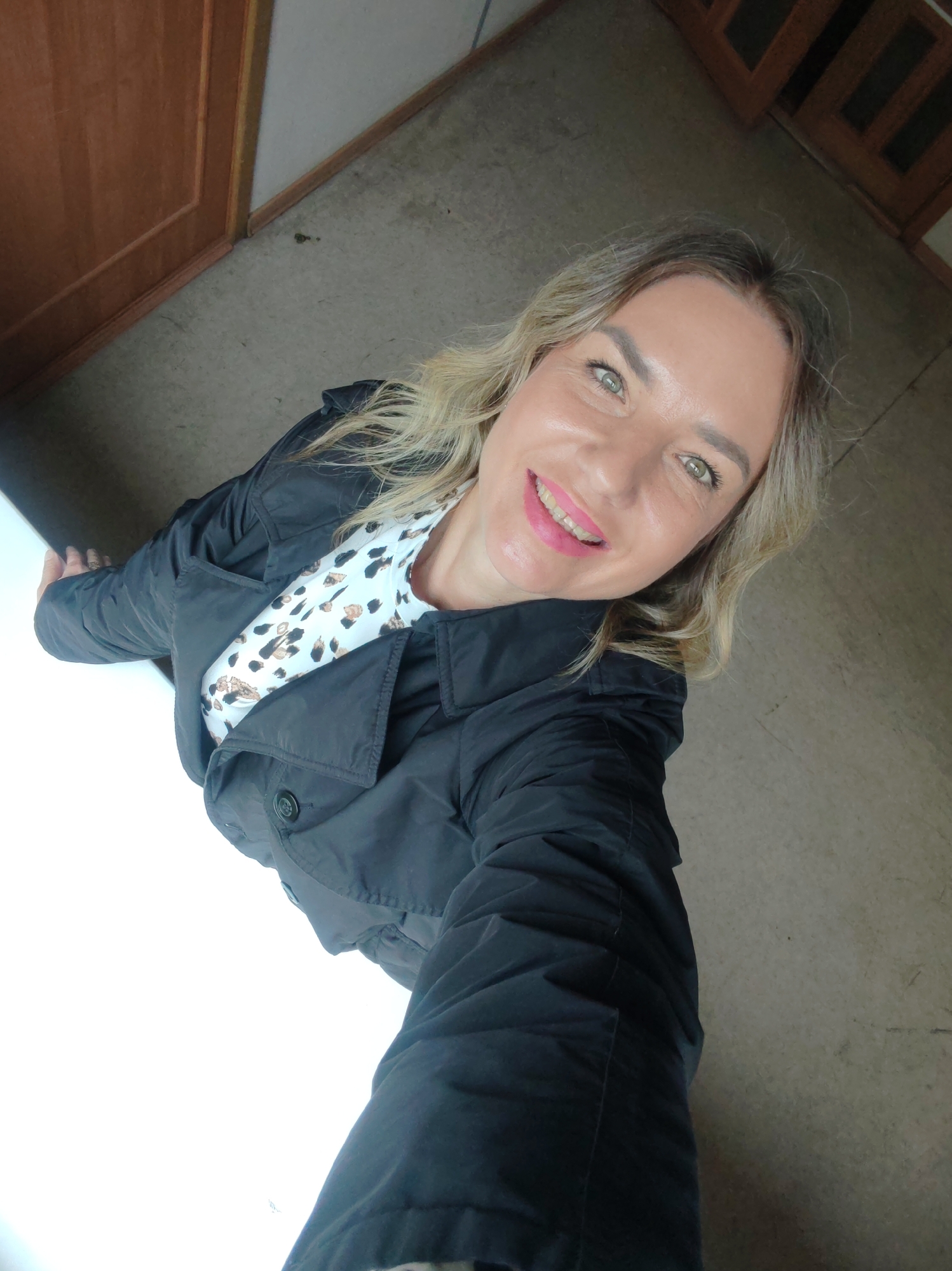 A woman with blonde hair, green eyes, and pink lipstick is smiling and looking directly at the camera, slightly angled from below. She is wearing a black jacket over a white shirt with a black and brown leopard print pattern. The background includes a light grey floor and a brown door frame. The location cannot be determined as Petropavlovsk-Kamchatsky based on the provided image.
