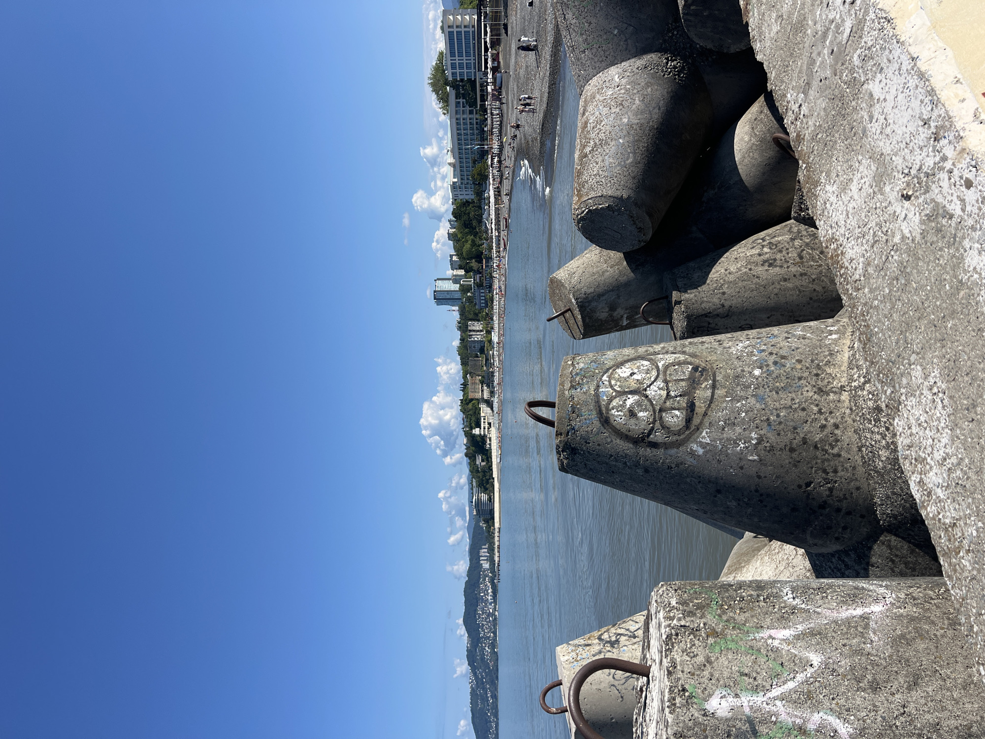 This is a photograph taken from a concrete breakwater overlooking the Black Sea coast in Sochi, Russia. The foreground is dominated by large, weathered concrete tetrapods, some of which are tagged with graffiti. Beyond the breakwater, the calm, gray-blue water of the sea stretches towards a pebble beach. Several people are visible on the beach, some lounging under umbrellas, others walking along the shore. Behind the beach, a line of white buildings, including hotels and resorts, stretches along the coastline, with more buildings and greenery climbing the hills in the background. The sky is a clear, bright blue with a few scattered white clouds. The overall scene depicts a sunny day at a popular seaside resort.