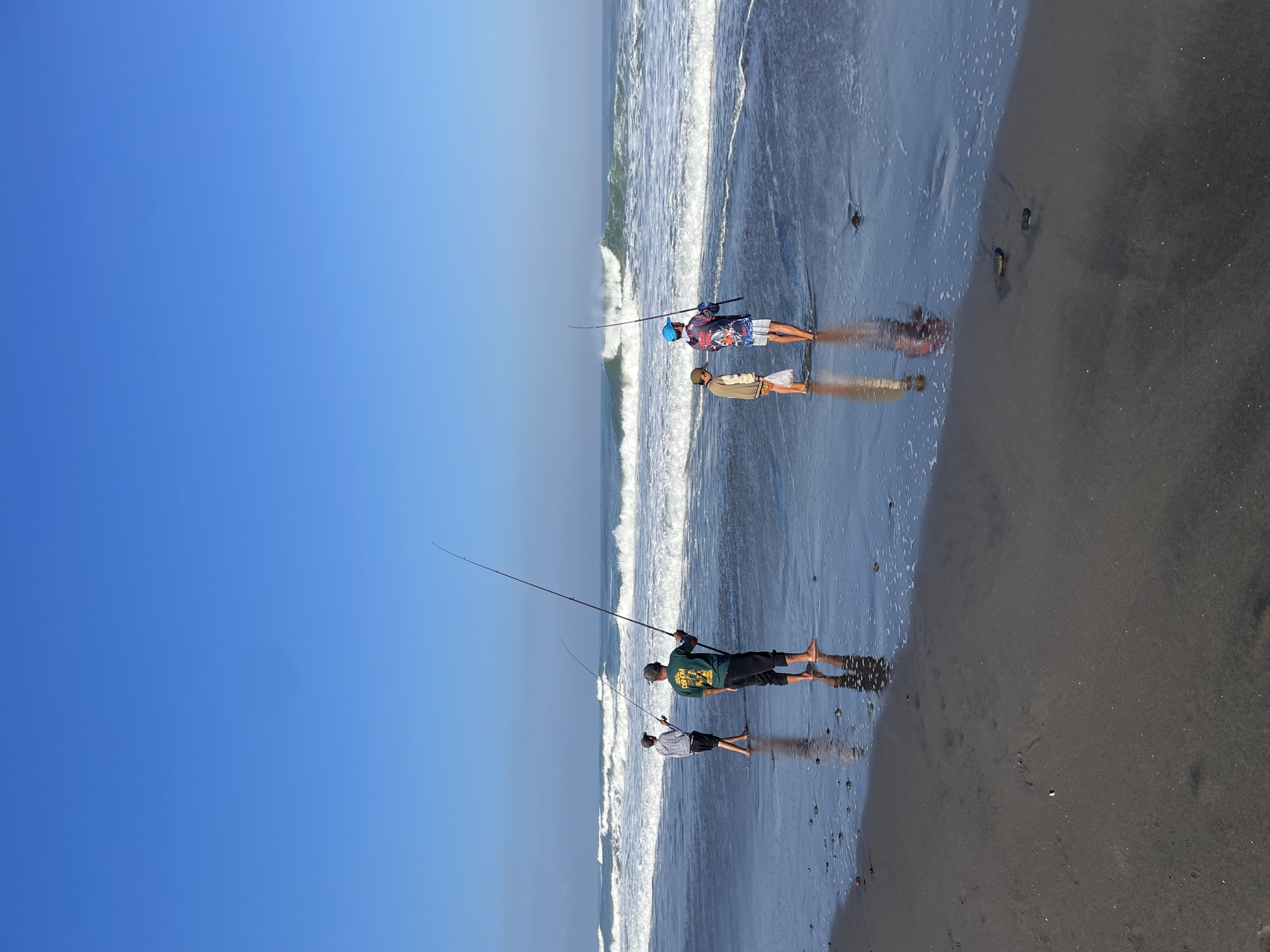 In Kediri, Indonesia, on a clear day, four individuals are seen fishing on a dark sand beach. Two adults and two children stand at the water's edge, holding fishing rods. The waves are moderate, creating white foam as they reach the shore. The sky is a vibrant blue, and the sun casts reflections on the wet sand. The scene captures a peaceful moment of recreational activity by the sea.