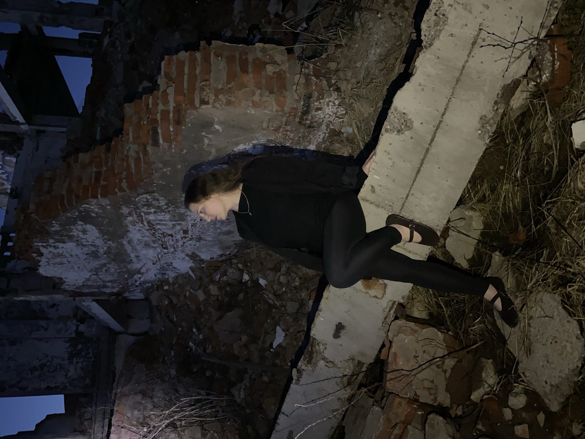 The image shows a woman lying on a concrete slab in an apparent ruined structure in Марковский, Russia. The woman is wearing a black shirt, black pants, and dark shoes with straps. The structure surrounding her is made of brick and concrete, with signs of decay. The setting appears to be at night.