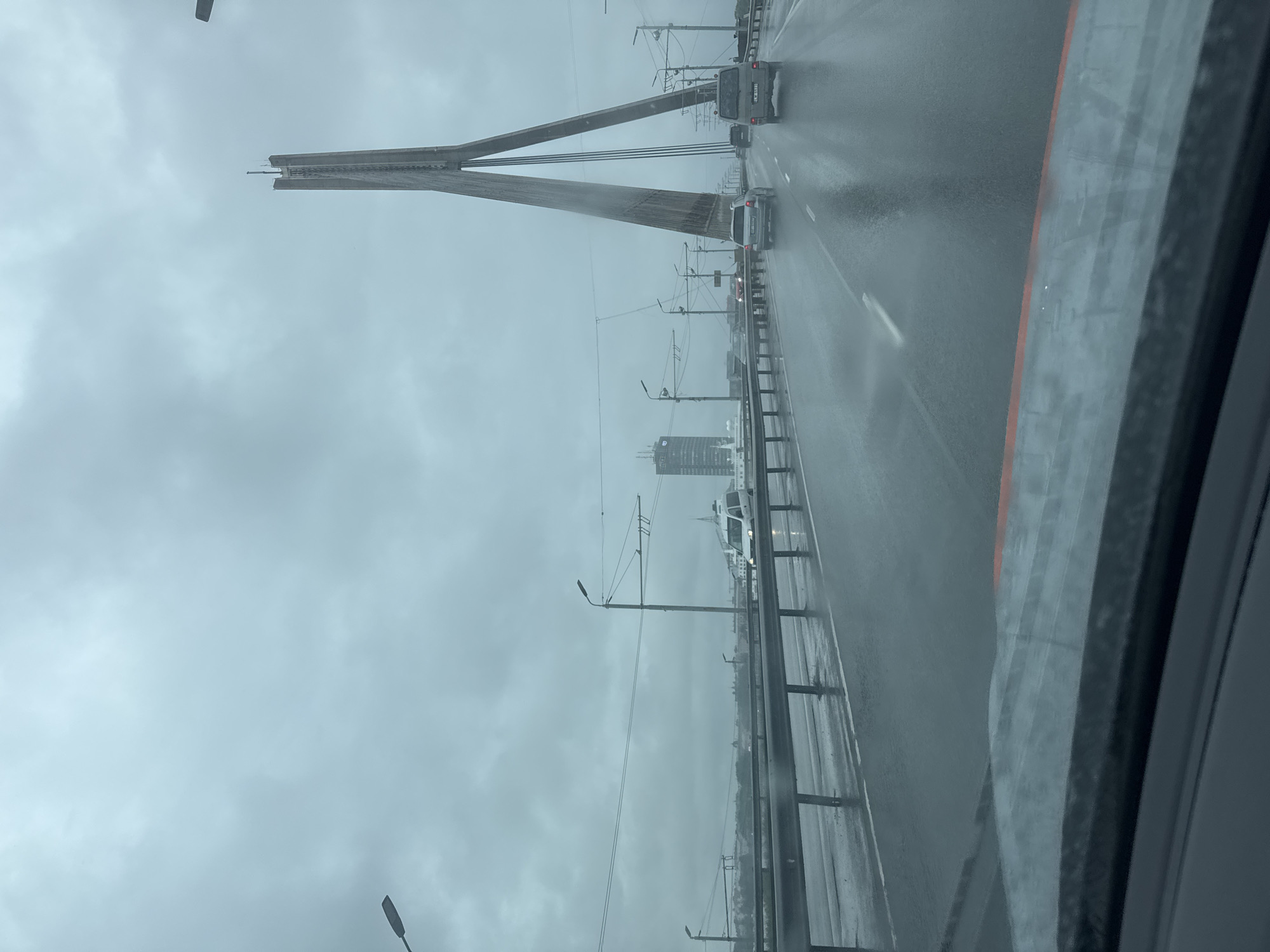 The image captures a view from a vehicle on a rainy day, traveling across a bridge in Riga, Latvia. The scene depicts a wet, overcast sky with heavy cloud cover. The bridge appears to be a cable-stayed bridge, with a prominent, tall, V-shaped pylon dominating the right side of the frame. The roadway is slick with rain, reflecting the ambient light and the headlights of the vehicles. Several cars are visible on the bridge, moving in the same direction as the viewer. A white van is in the foreground on the left lane, and other vehicles are further ahead. In the distance, a tall, modern building with a distinctive architectural design is visible on the left side of the frame, alongside other structures that suggest an urban landscape. The overall atmosphere is somber and indicative of inclement weather. The presence of streetlights and overhead wires further clarifies the setting as a public roadway.