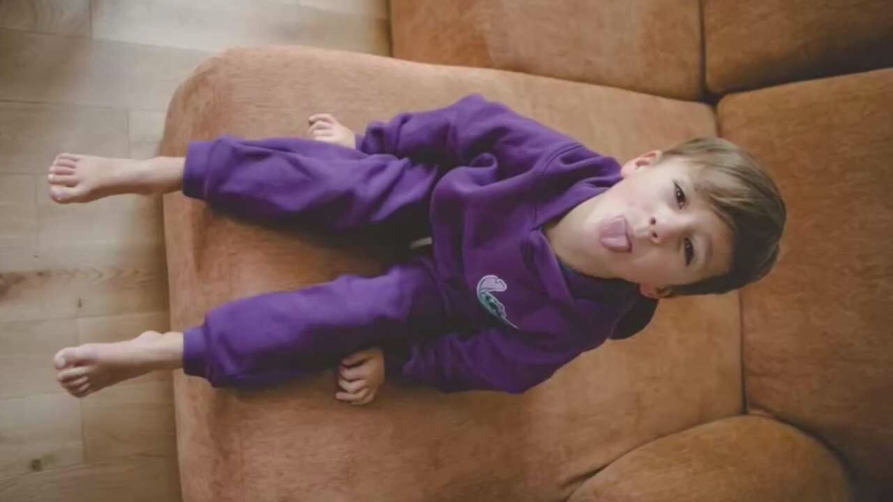 A young Caucasian boy, with light brown hair, is sitting on a brown couch with his legs hanging off the edge. He is wearing a purple hooded sweatshirt with a light blue graphic on the left chest and matching purple sweatpants. The boy is sticking his tongue out at the camera. His bare feet are visible, with his toes spread apart. The floor beneath the couch is made of light-colored wood planks. The background is blurred, but appears to be an indoor setting with the couch occupying most of the frame.