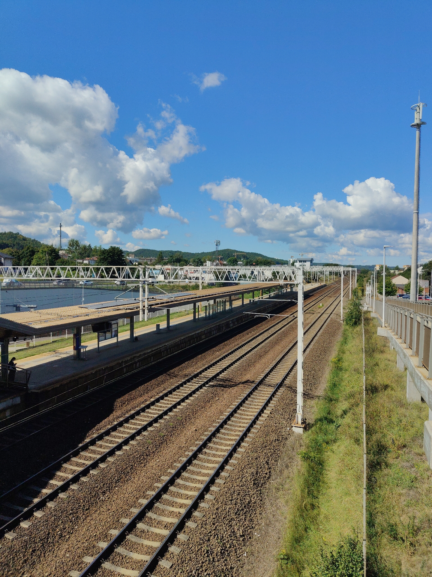 The image shows a train station with multiple railway tracks leading into the distance. The tracks are surrounded by gravel and vegetation. A covered platform with benches and signage is visible to the left, with a person standing on it. Overhead power lines are supported by white poles. In the background, there are buildings, trees, and hills. The sky is blue with scattered white clouds. This is the train station in Rumia, Poland.