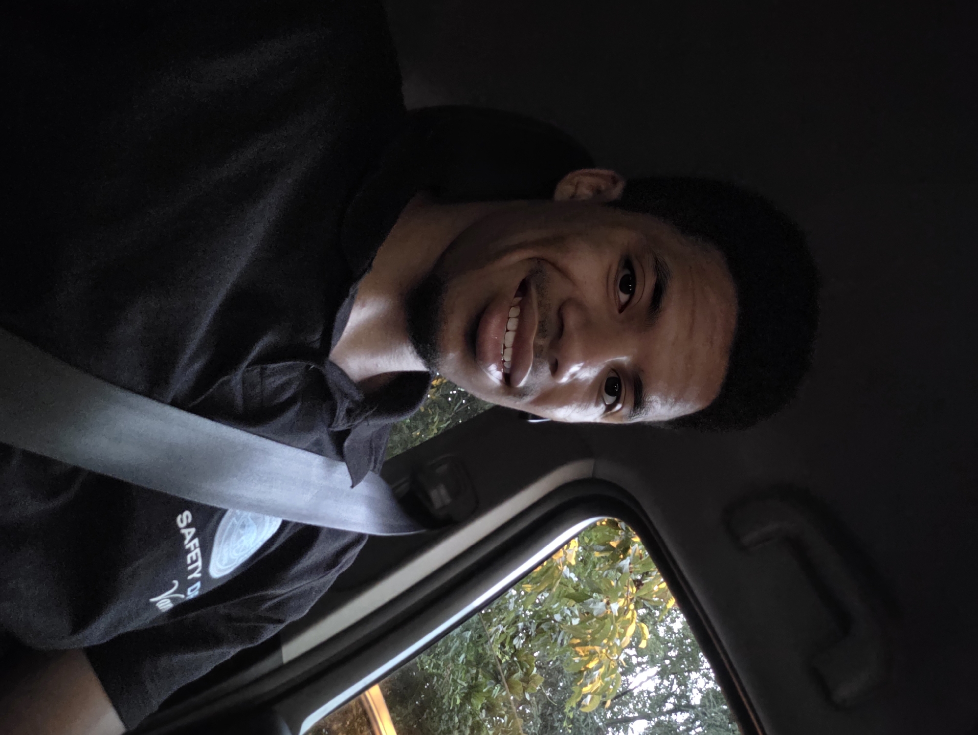 A man is captured in a selfie from inside a vehicle. He has dark skin and short, dark hair. He is wearing a black polo shirt with a logo that appears to say "SAFETY D" and "Van" on it. A silver seatbelt is visible across his chest. The background shows foliage through a car window, suggesting an outdoor setting. The car interior is dark, with a handle visible on the ceiling. There are no explicit indicators of the city of Varna, Bulgaria in the image.