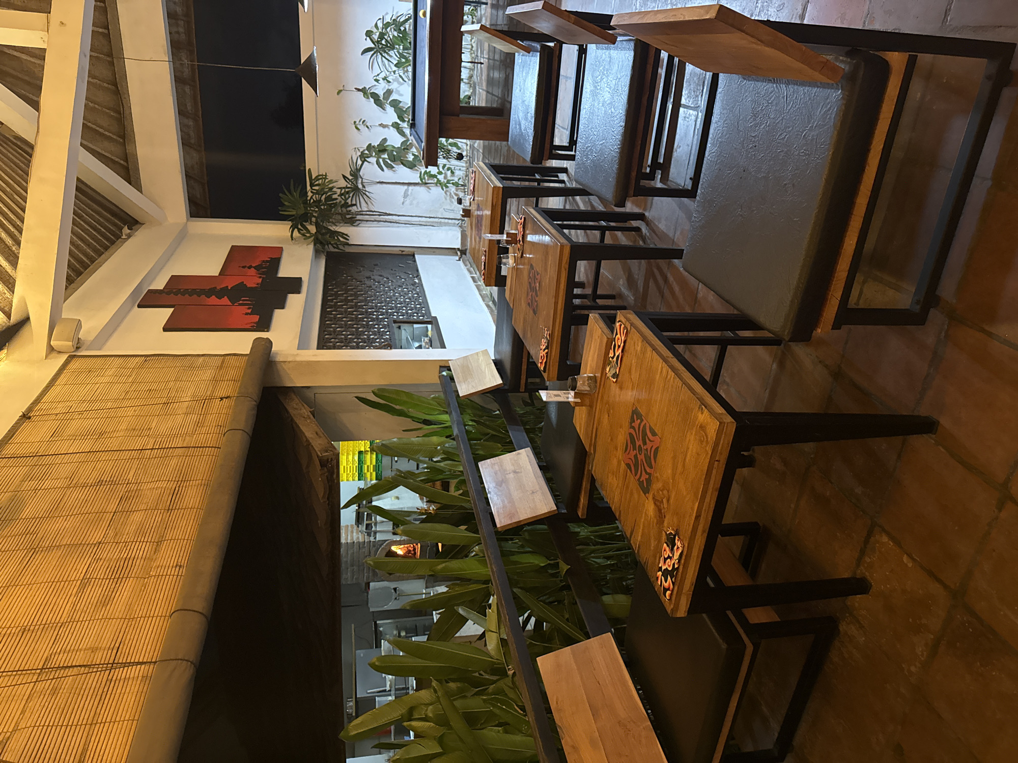 The photograph depicts a restaurant setting. Tables with wooden tops and dark frames are arranged in rows. Some tables have seating with padded cushions. There are plants decorating the space, with some positioned behind a metal railing. Outside, through an opening, a glimpse of a pizza oven can be seen. Above, a section of the ceiling has bamboo thatching. A wall is decorated with a silhouette design in red and black. The location is Unknown, Unknown.
