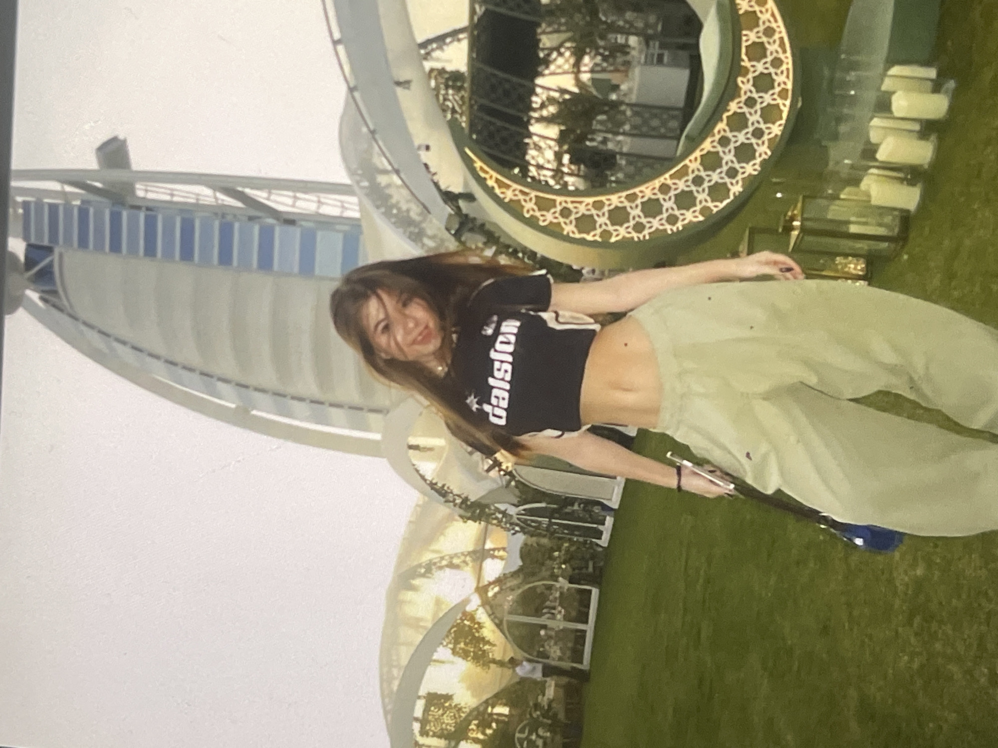 The image features a young woman posing outdoors, smiling towards the camera. She has long, straight brown hair and is wearing a black, short-sleeved crop top with the word "dalsion" visible in white text across the front. Her attire is completed by loose-fitting, light green or khaki pants. She holds a smartphone in her left hand.
The background is dominated by a distinctive, sail-shaped building with a white and light blue striped facade, strongly resembling the Burj Al Arab in Dubai. To the right of the woman, a large, decorative crescent moon-shaped structure with intricate golden patterns is prominently displayed, alongside several rectangular lamps arranged on the ground. The foreground consists of lush green grass, suggesting a well-maintained lawn or garden area. In the distance, other outdoor structures, possibly pergolas or seating areas, are visible under a bright, clear sky.
The scene takes place during daylight hours, with ample natural light indicating bright, clear weather. The woman appears happy and relaxed while posing.
The provided location context for this image is Poltava, Ukraine; however, the visual elements, particularly the iconic sail-shaped building and crescent decoration, are strongly indicative of a Middle Eastern setting, specifically Dubai.