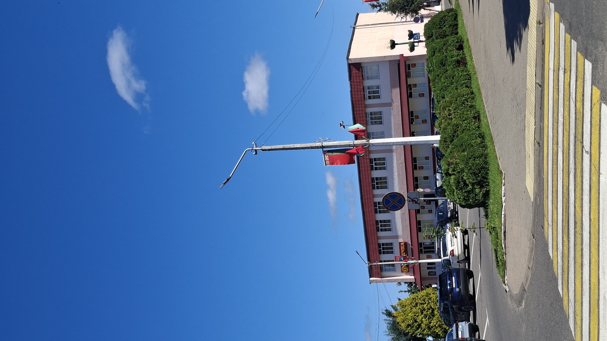 In Myadzyel, Belarus, a bright, clear blue sky with a single small white cloud dominates the upper portion of the image. Below the sky, a modern building with a red tiled roof and light-colored walls is visible. The building features a row of evenly spaced windows across its facade. In front of the building, a well-maintained row of green bushes lines a sidewalk. A street with a pedestrian crosswalk marked by yellow and white stripes is in the foreground. Several cars are parked or driving on the street. A lamppost with a light fixture and flags attached stands prominently on the right side of the street, near the building. There are also traffic signs visible. The overall scene depicts a calm day in a town setting.