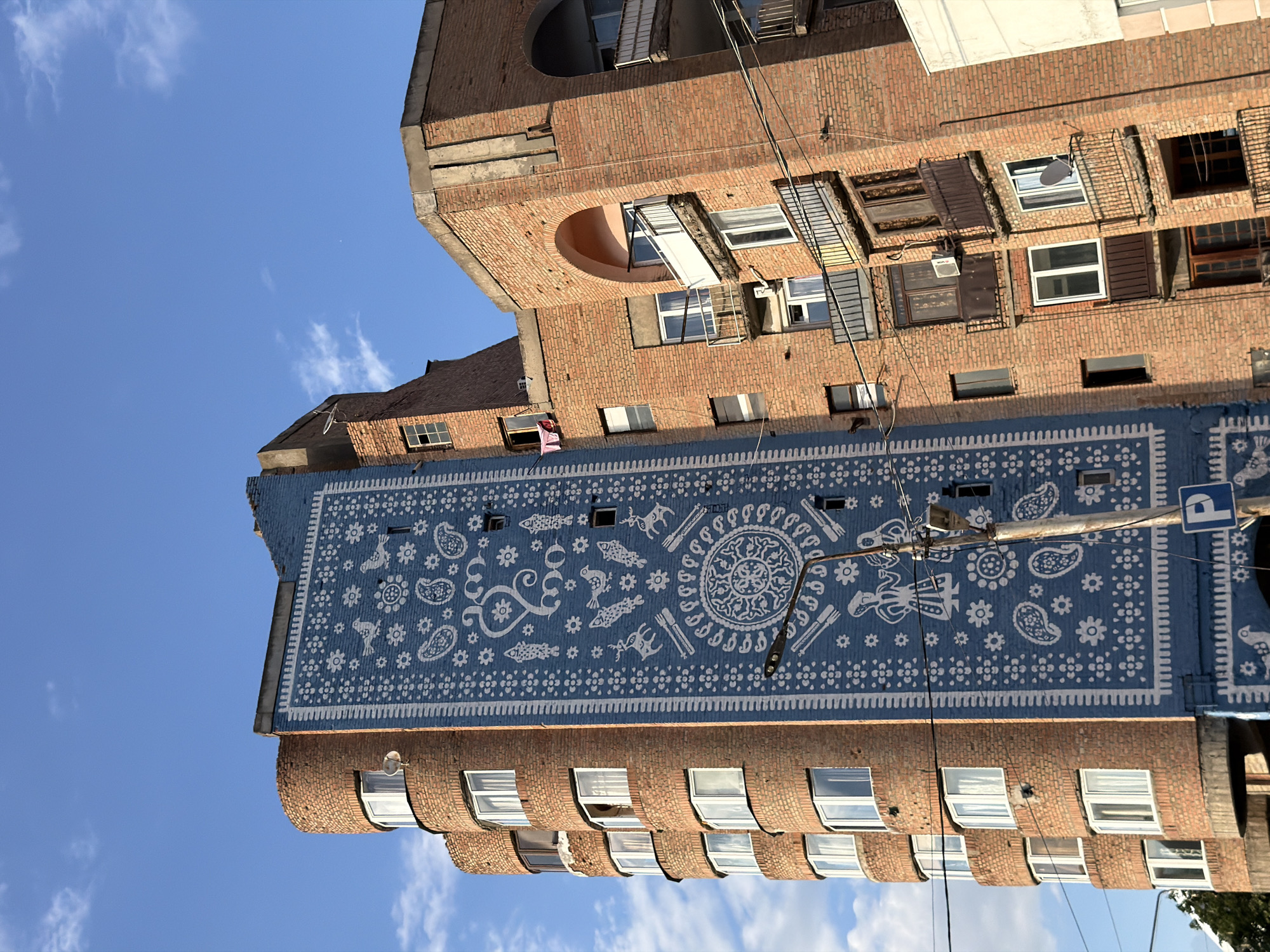 The image shows a residential building in Gori, Georgia, with a large blue mural painted on one side. The mural is decorated with white patterns, including paisleys, flowers, birds, and figures resembling people in traditional attire. The building itself is made of reddish-brown brick and has a curved section with multiple windows. To the right of the muraled section, another part of the building is visible, also constructed of brick and featuring balconies with wrought-iron railings and arched window openings. Several electrical wires are strung across the scene, and a lamppost with a parking sign is situated in front of the mural. The sky above is blue with scattered white clouds.
