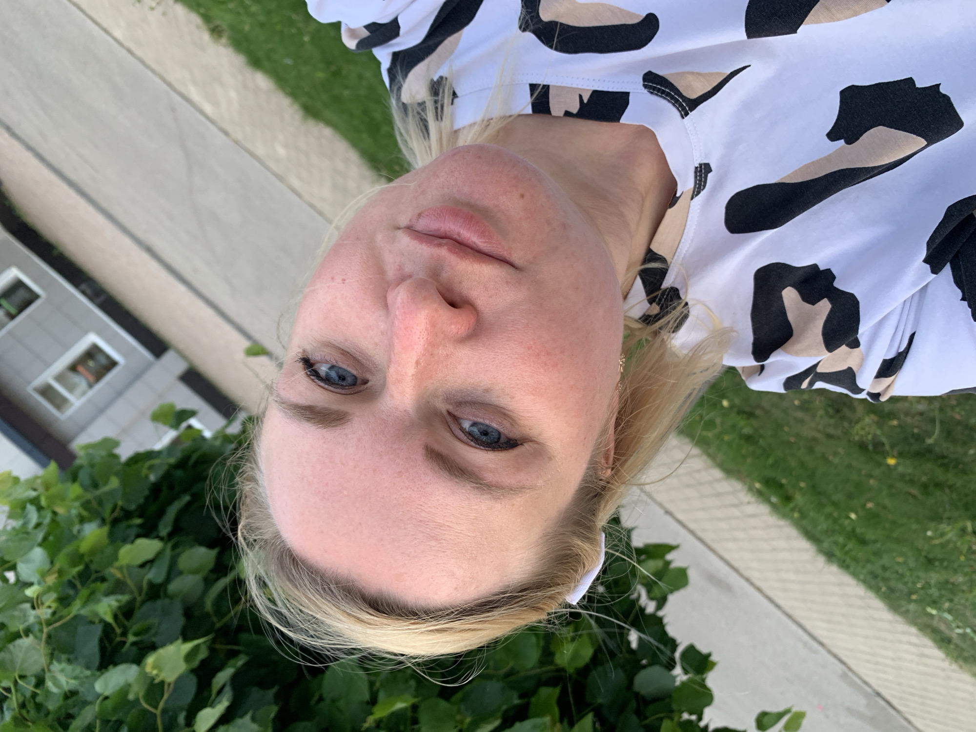 A woman with fair skin and blonde hair is facing the camera with a slight tilt of her head. Her eyes are light blue. She is wearing a black and white patterned shirt. The background includes green foliage in the lower portion, and concrete with a grassy area to the side. A building is partially visible in the upper left. There is no evidence to confirm the location is Chaikovsky, Russia.